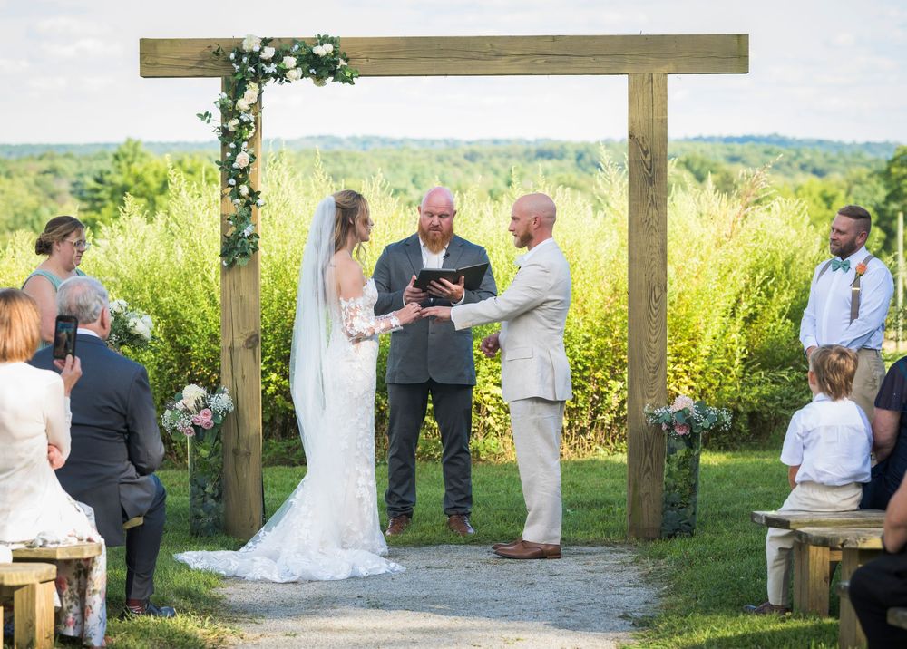 An outdoor wedding ceremony takes place under a wooden arch with rolling hills and wildflowers in the background.