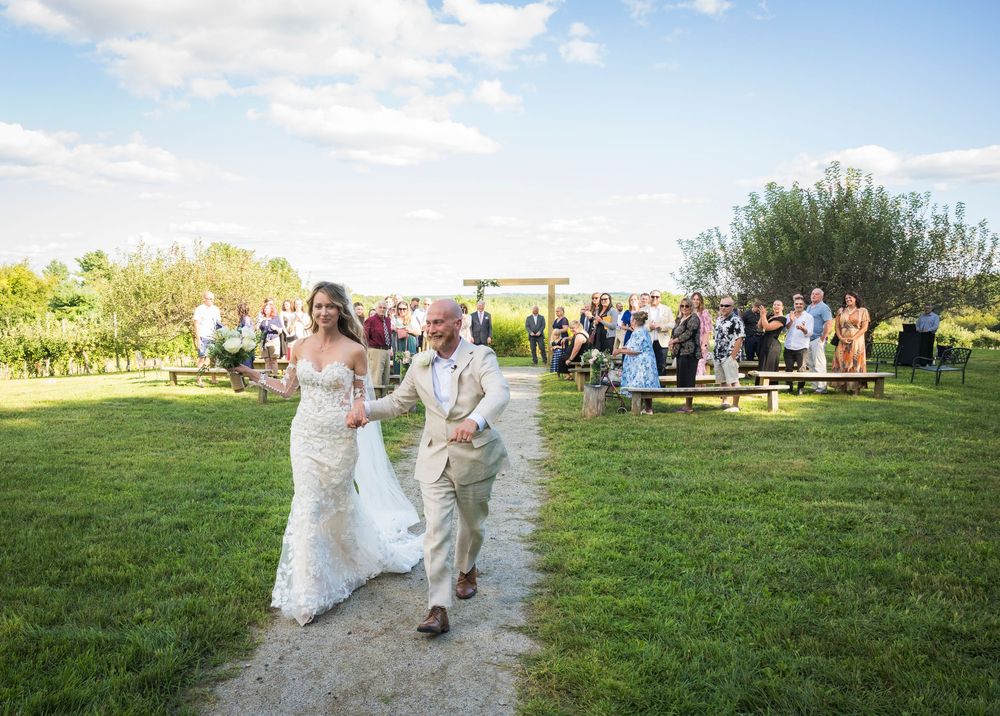 Wedding guests seated on rustic benches during an outdoor ceremony surrounded by green fields and blue skies.