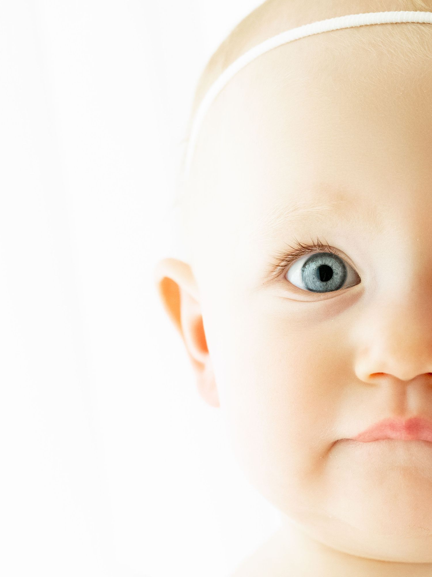 Close up view of a baby's face showing blue eyes, a delicate nose, and soft skin against a neutral background.