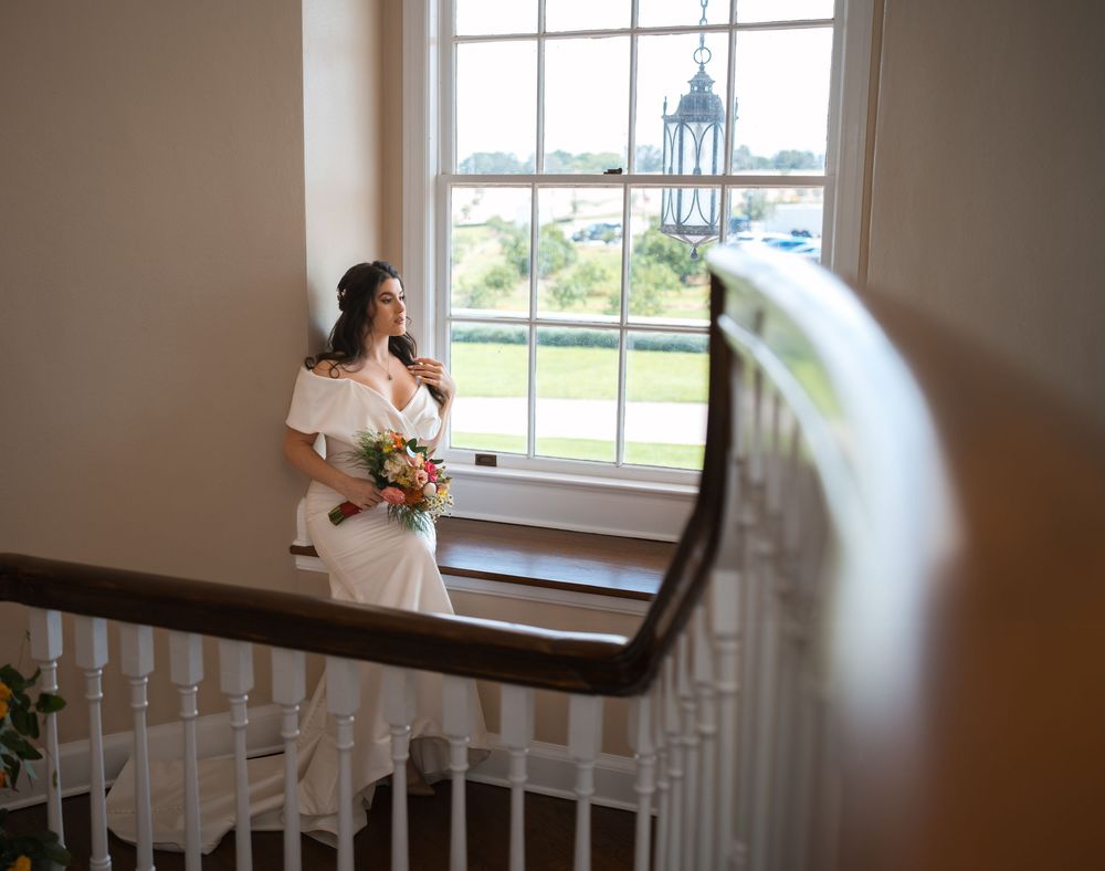 Someone holding a bridal bouquet stands by a window on a staircase landing in an elegant interior setting.