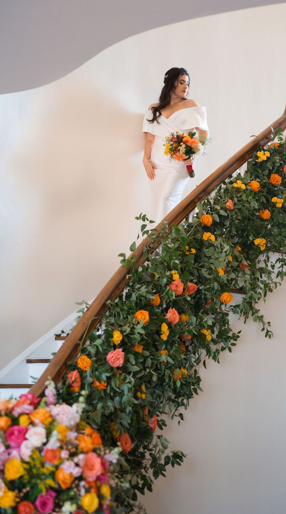 A bride in a white off-shoulder gown descends an elegant staircase decorated with vibrant orange and pink floral arrangements.