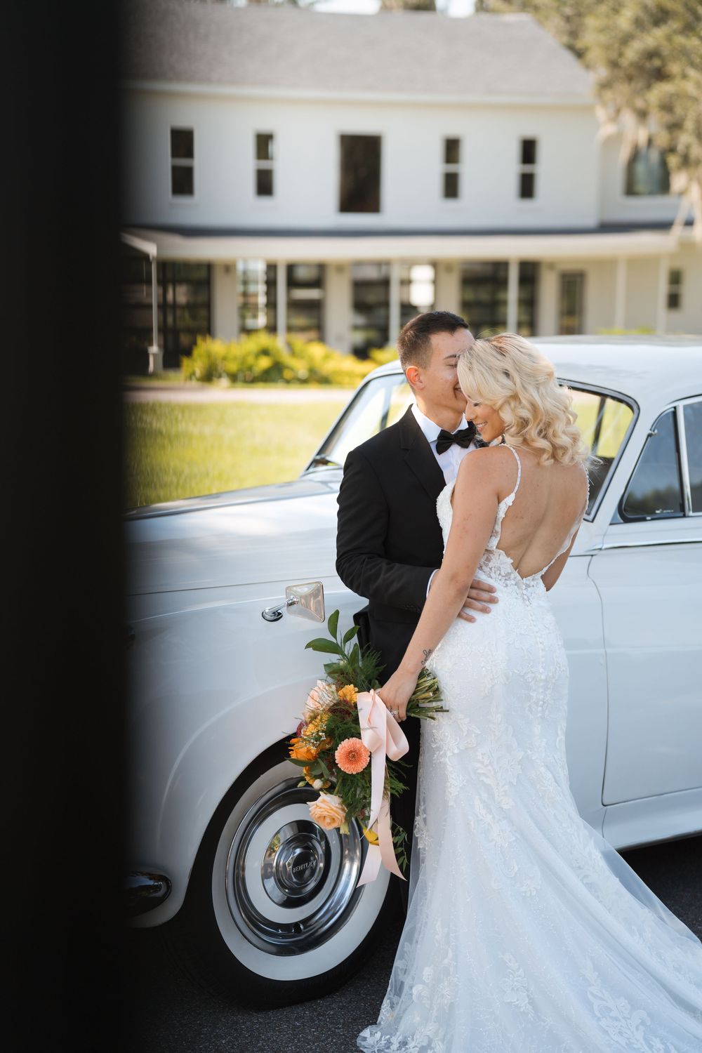 A newlywed couple poses romantically next to a classic white Rolls Royce on their wedding day.