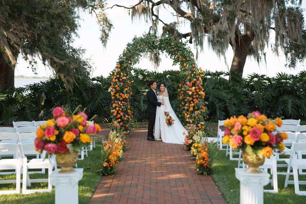 Outdoor garden wedding ceremony setup with orange floral arch and white chairs on brick path under Spanish moss trees.
