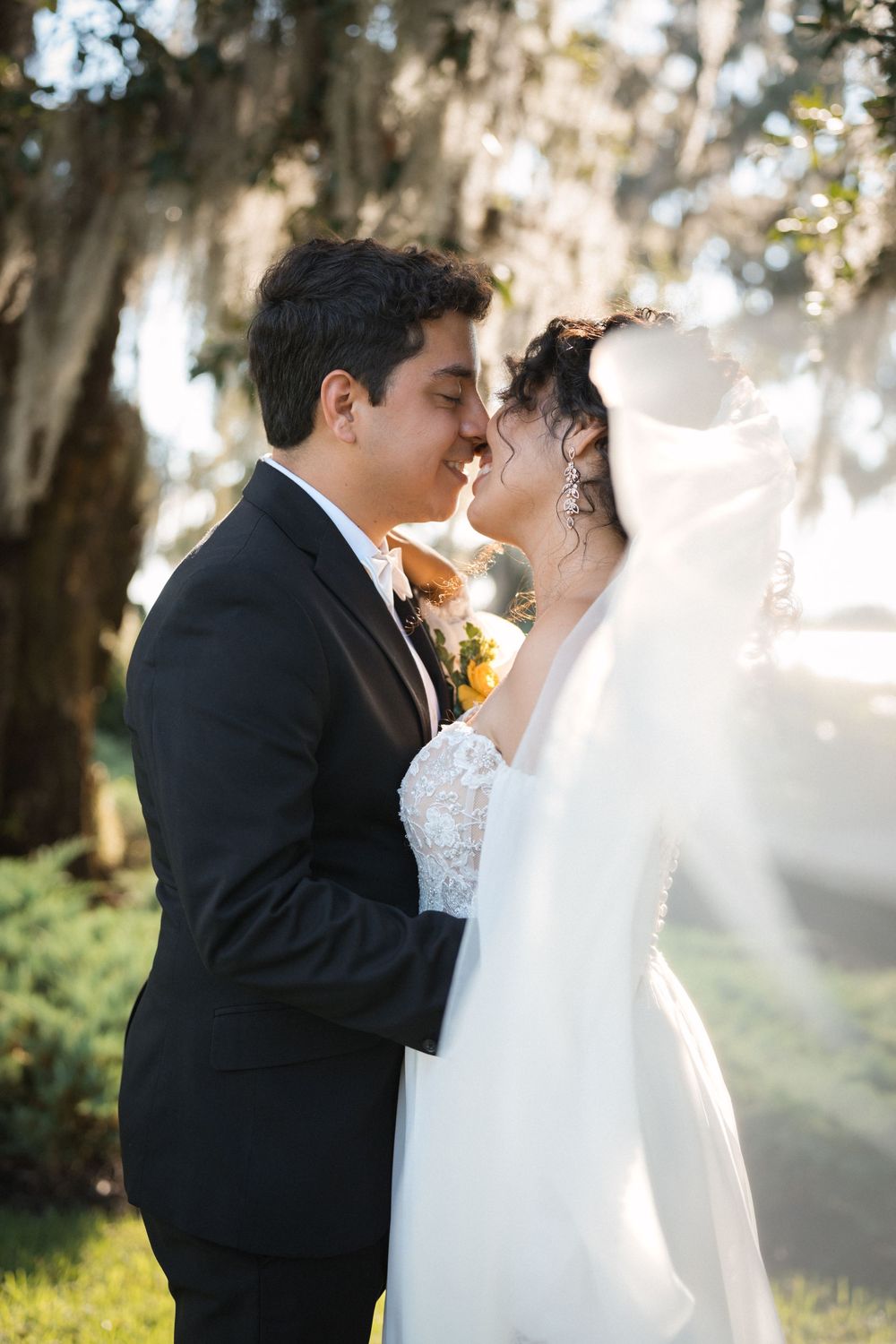 Newlywed couple shares intimate moment outdoors with sunlight filtering through Spanish moss draped trees.