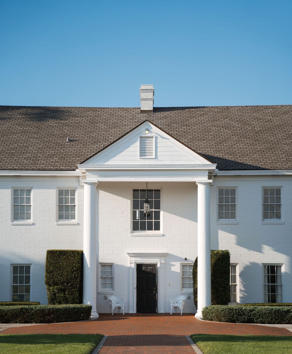 White colonial-style house with symmetrical windows, columns framing the entrance, and manicured hedges along a brick walkway.