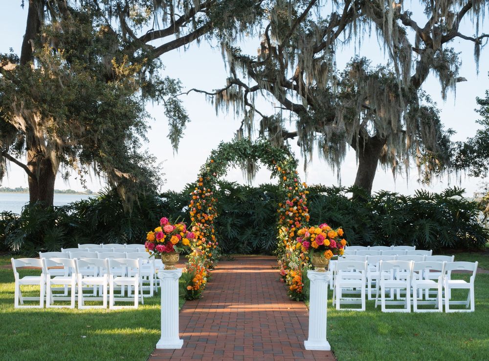 Outdoor wedding ceremony setup with white chairs and orange floral arrangements under moss-draped oak trees.