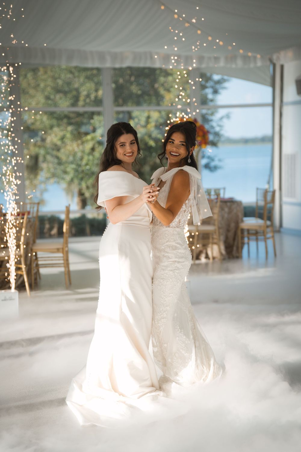 Two brides in elegant white wedding dresses dance together in a venue decorated with twinkling fairy lights.