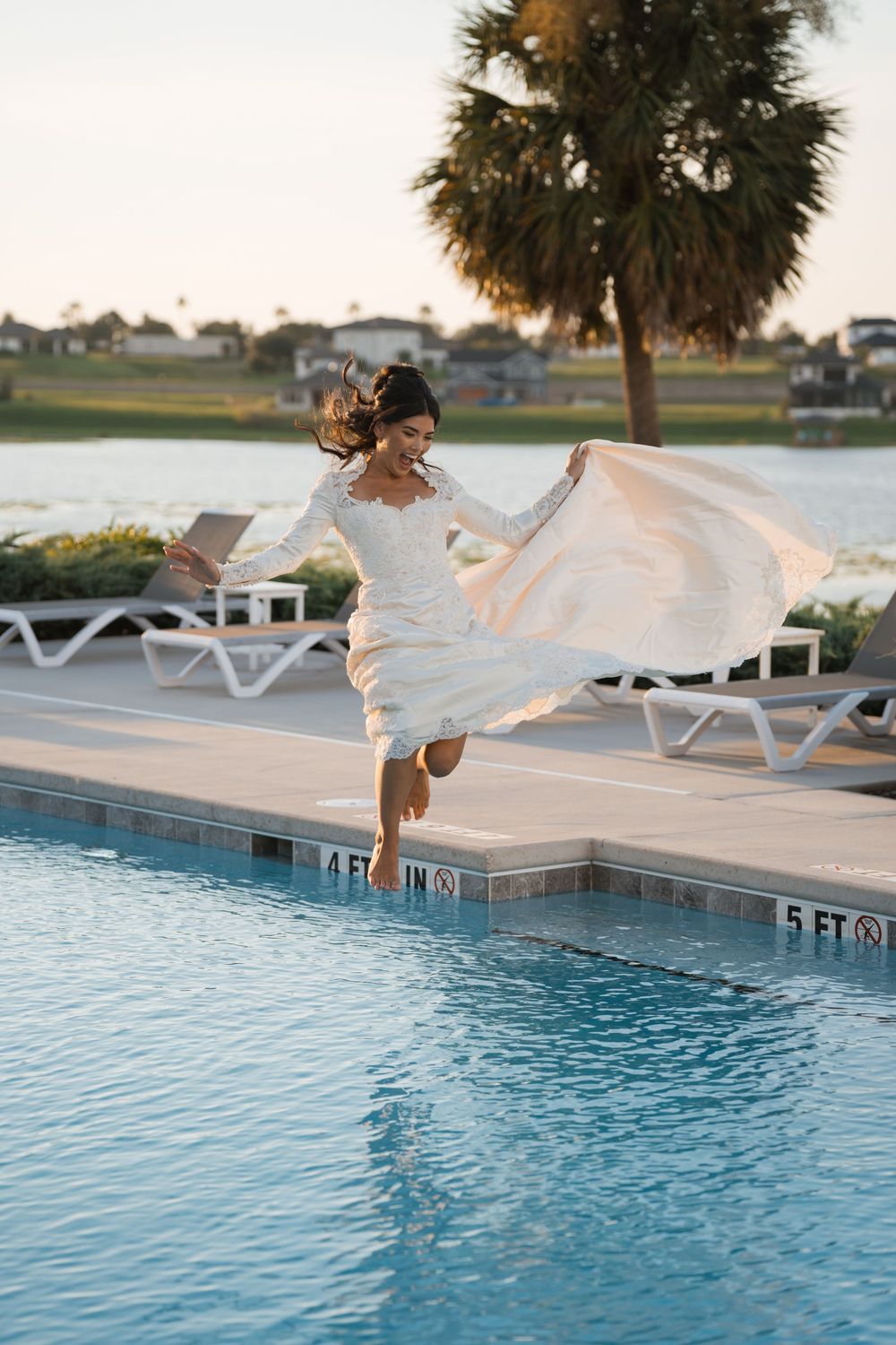 Figure in flowing white dress leaps gracefully into a poolside at sunset with palm trees in the background.