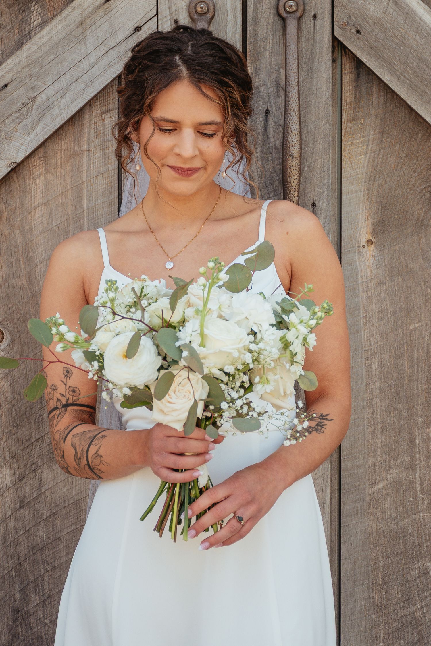 A bride in a white wedding dress holds a beautiful bouquet of white roses and greenery against a rustic wooden backdrop.