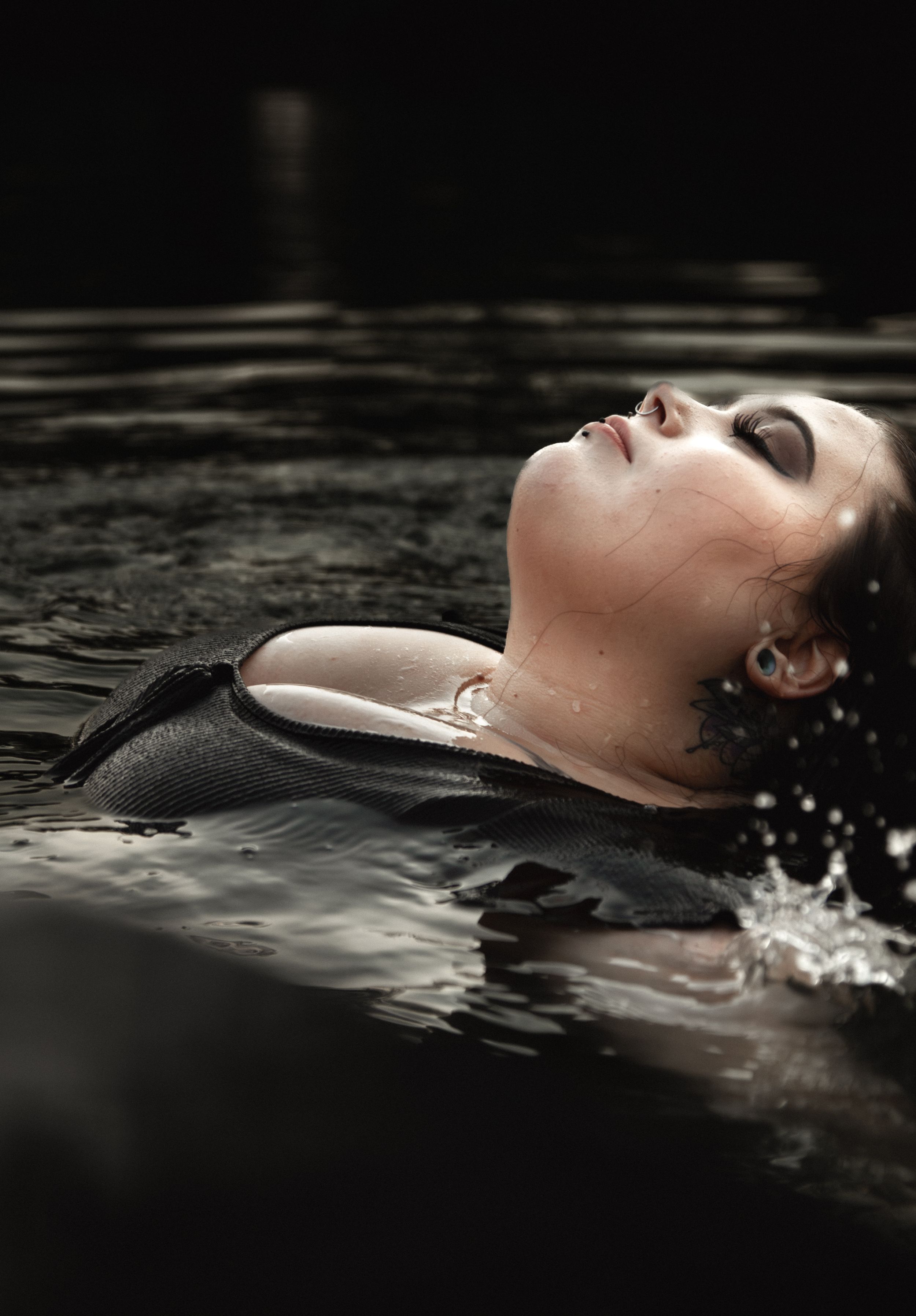 Person floating peacefully in dark water with closed eyes and wet hair, creating a serene and moody atmosphere.