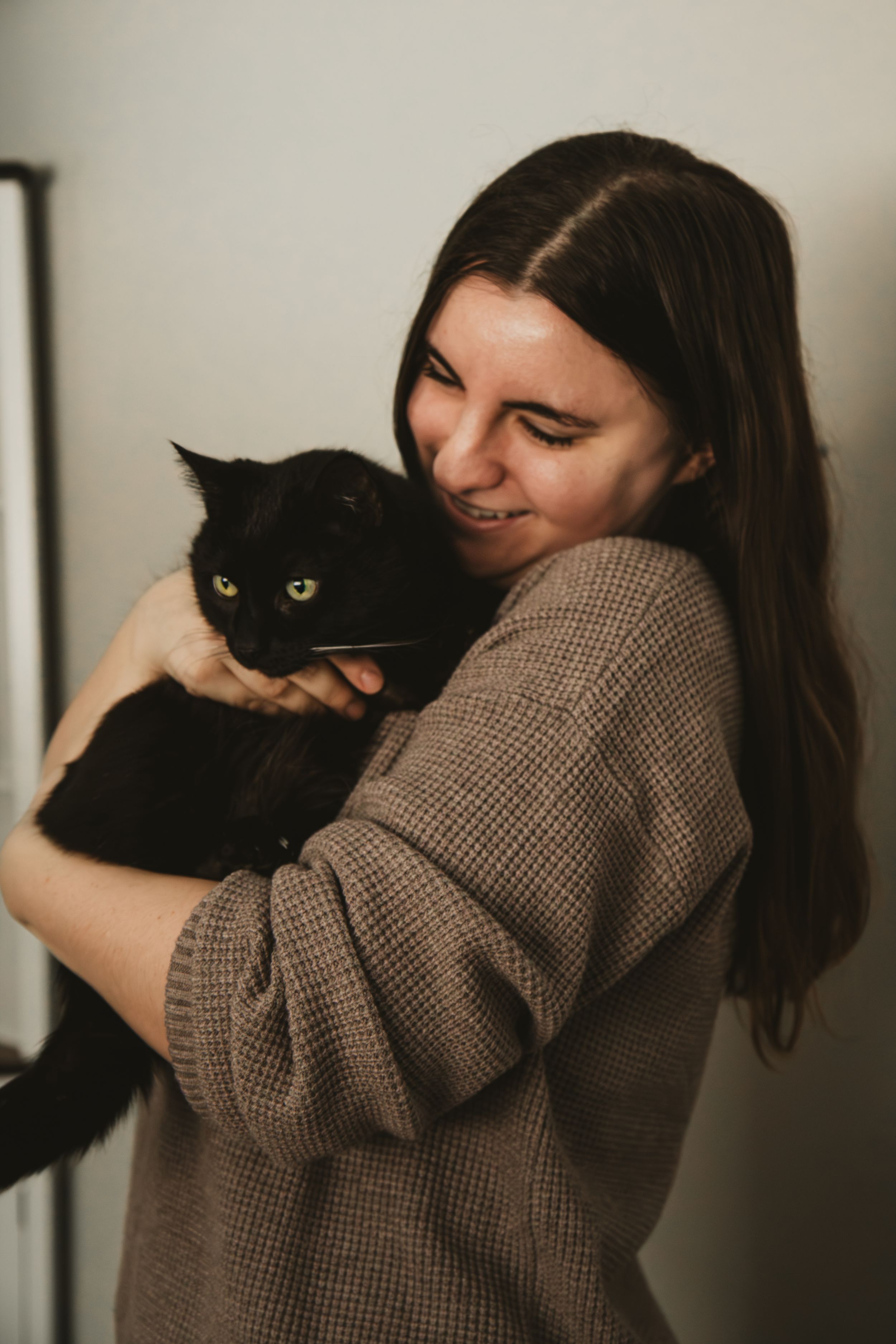 A person in a brown sweater hugs and smiles at their black cat with bright yellow eyes in a warm indoor setting.