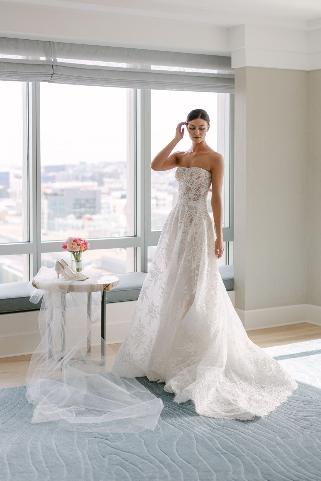 A strapless lace wedding gown with flowing train and delicate floral details stands in a bright high-rise apartment room.