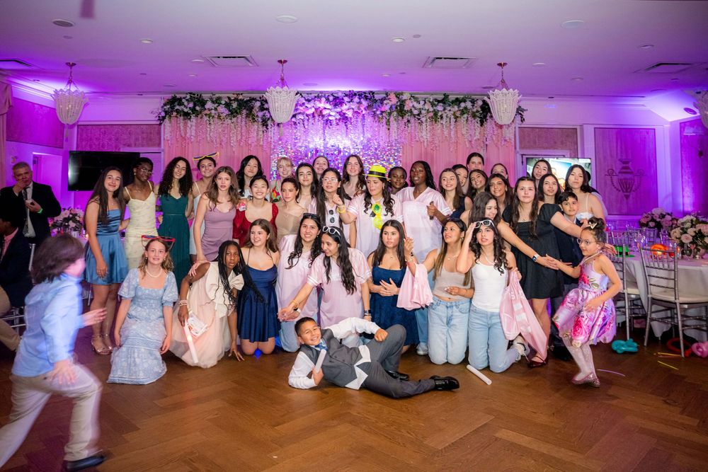 Large group celebrating at a wedding reception with purple uplighting and floral decor on dance floor.