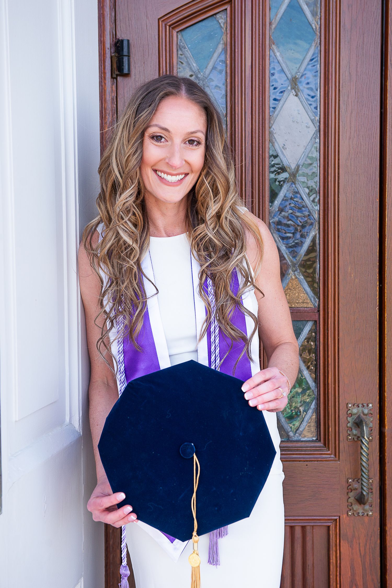 A person in a white top and navy blue skirt poses at a doorway with graduation regalia.
