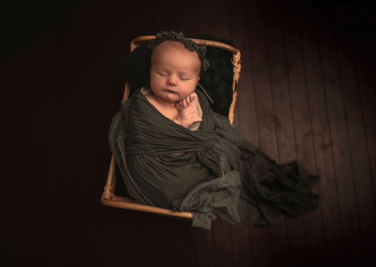 Sleeping newborn baby wrapped in dark green blanket laying in wooden basket against dark wood floor.