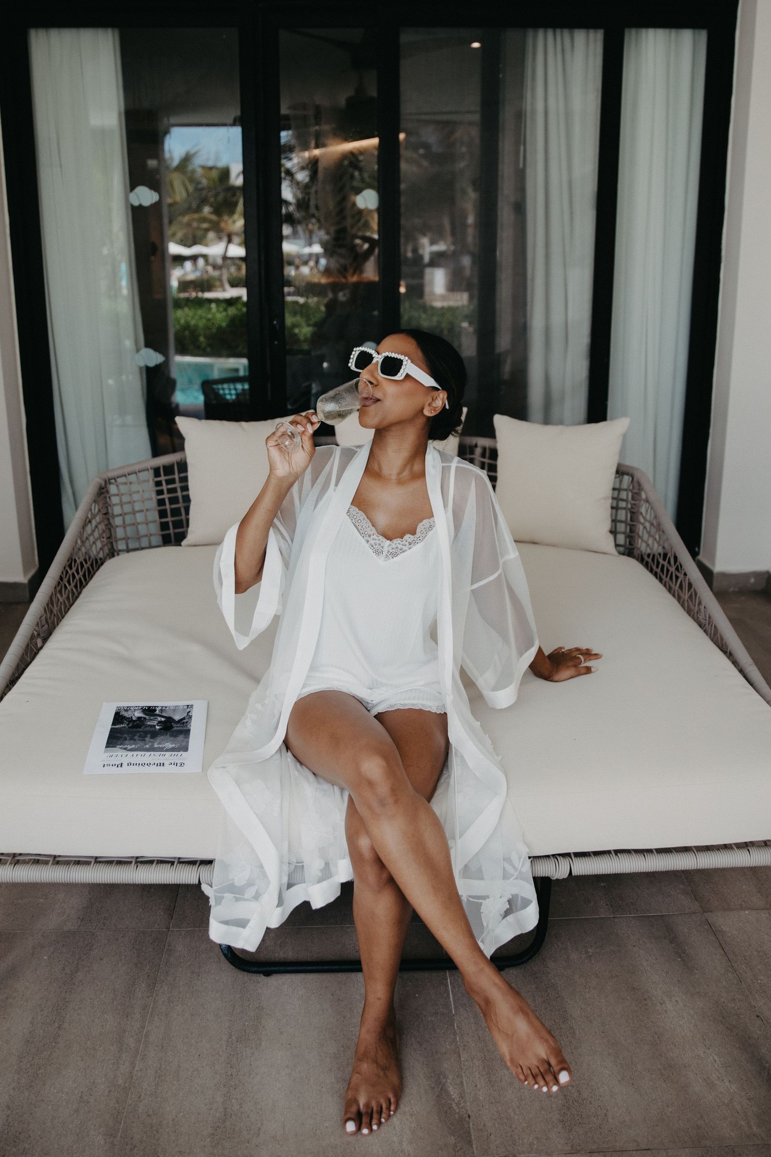 Person relaxing on outdoor daybed wearing white robe and sunglasses while sipping champagne.
