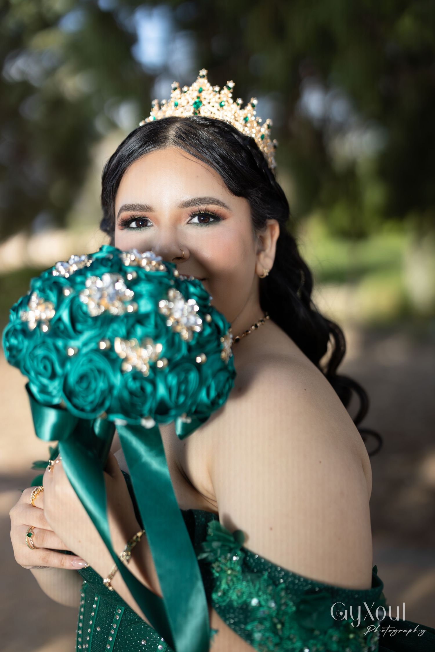 A teal quinceañera bouquet with crystal embellishments and satin ribbons is held by someone wearing a crown and teal gown.