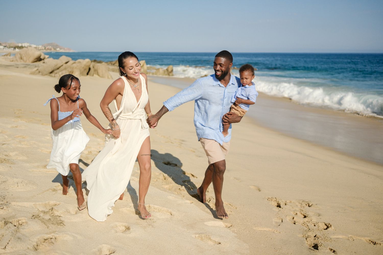 Six white-clad figures dance expressively on a beach at sunset casting long shadows in the sand.