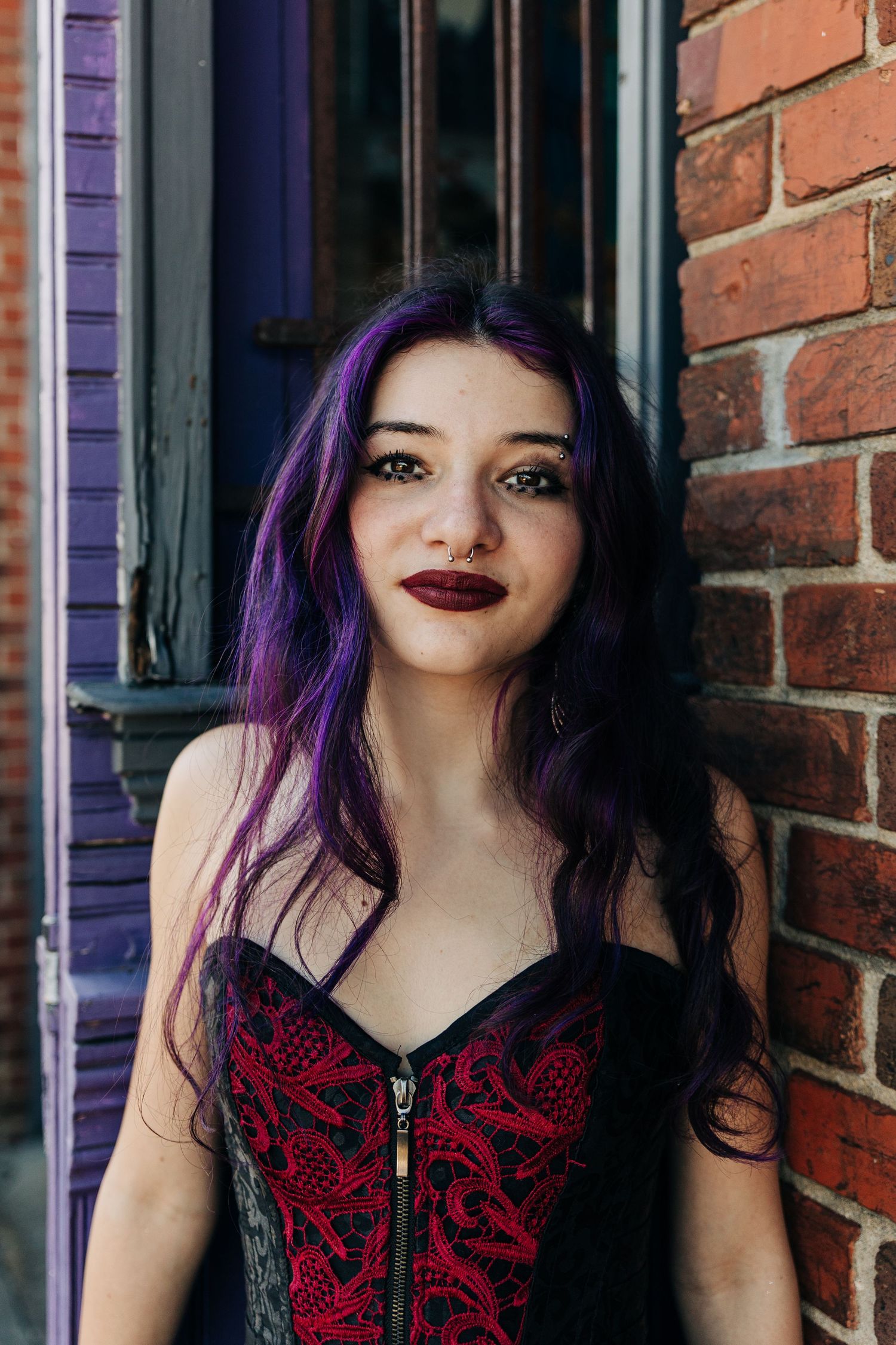 A person with vibrant purple hair wearing a red and black corset poses against a brick wall in Monroe, NC for senior photos.