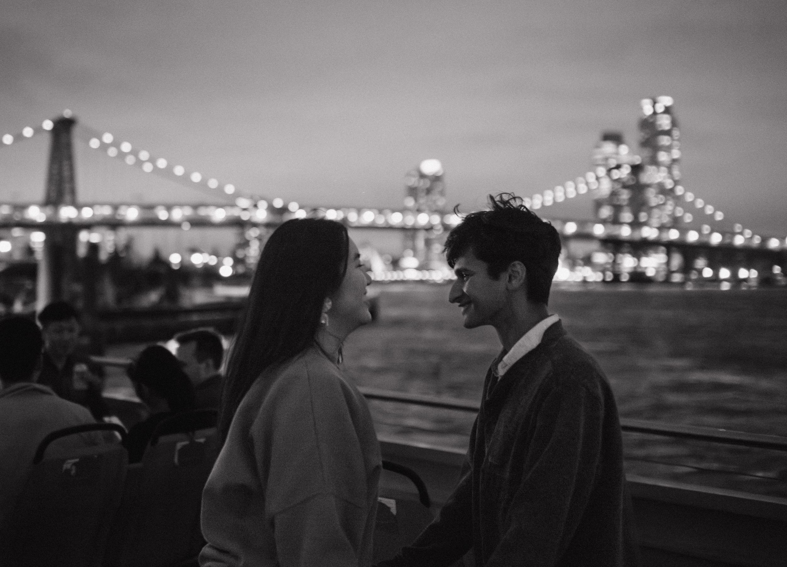 A romantic couple shares an intimate moment at night with the illuminated Brooklyn Bridge in the background.