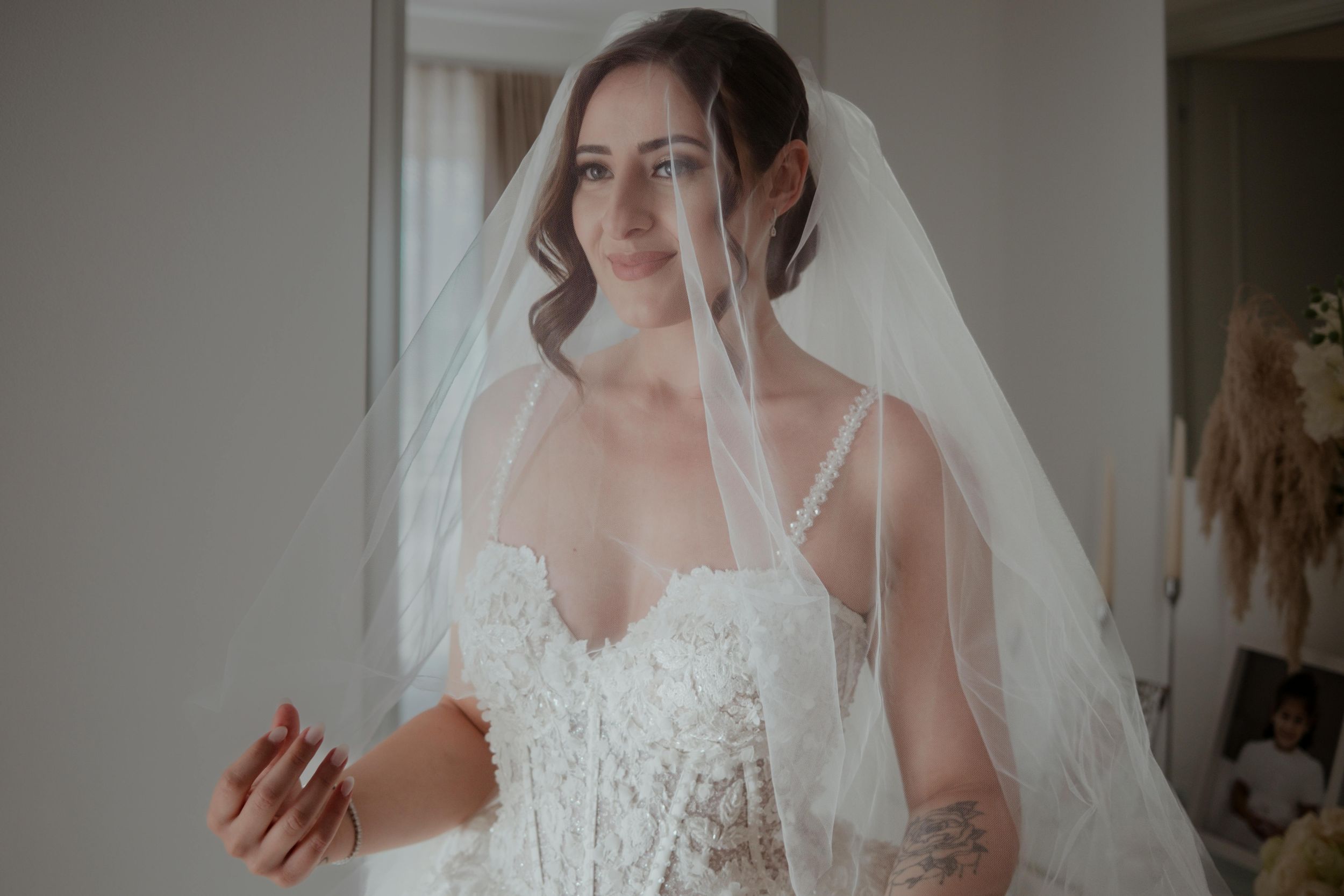 A bride in a lace wedding dress and veil stands in front of a window during her wedding day preparations.