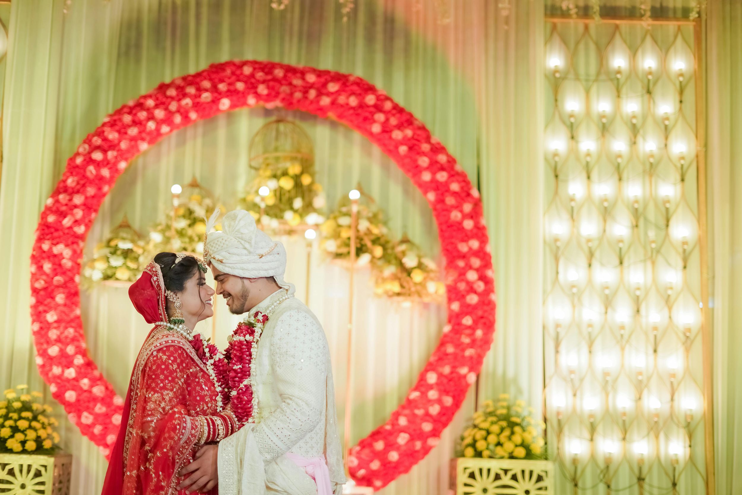 A wedding couple shares a romantic moment under a stunning red balloon arch at an elegantly lit venue.