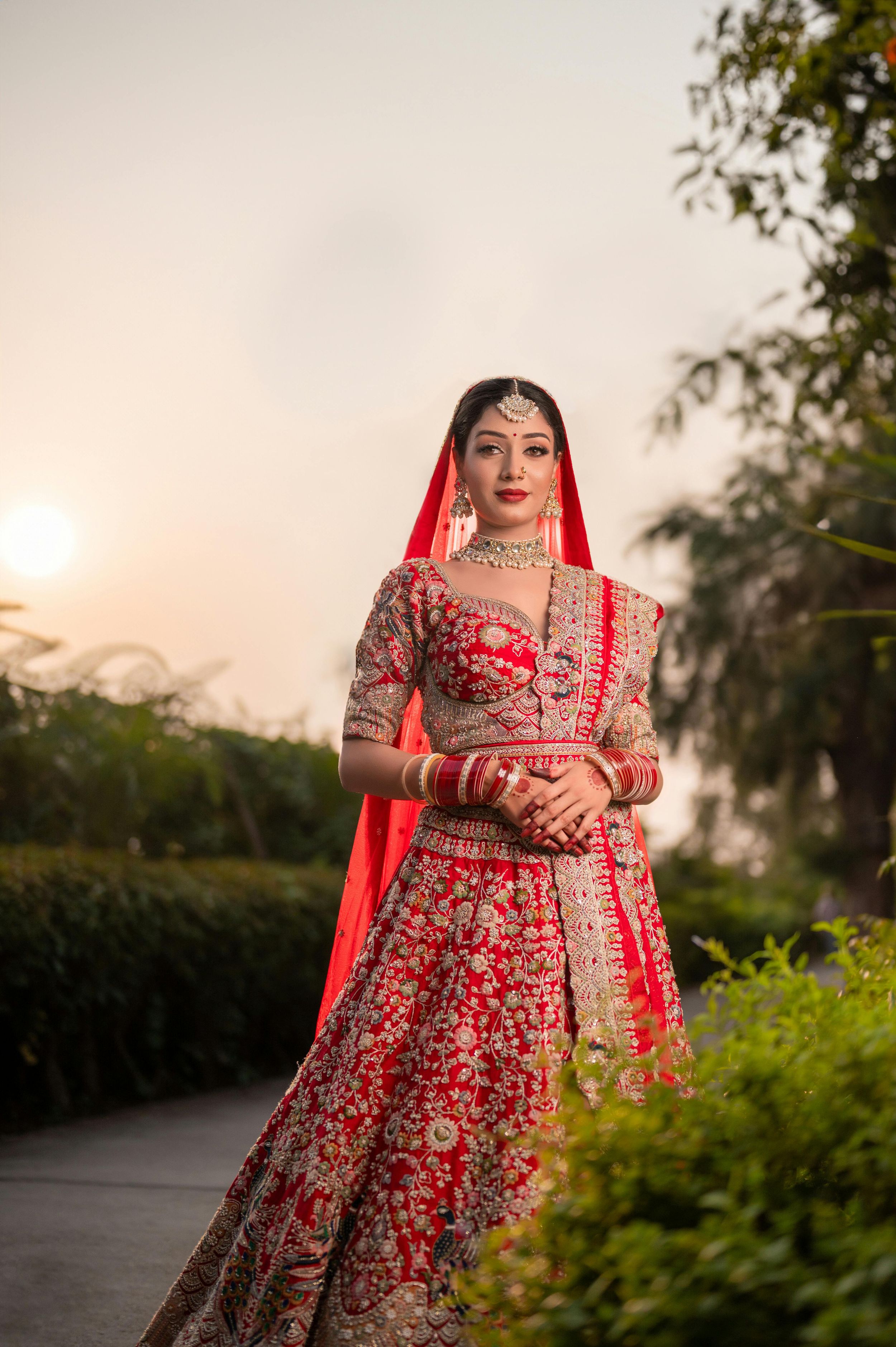 A bride wearing a stunning red and gold embroidered lehenga poses outdoors during sunset in a garden setting.