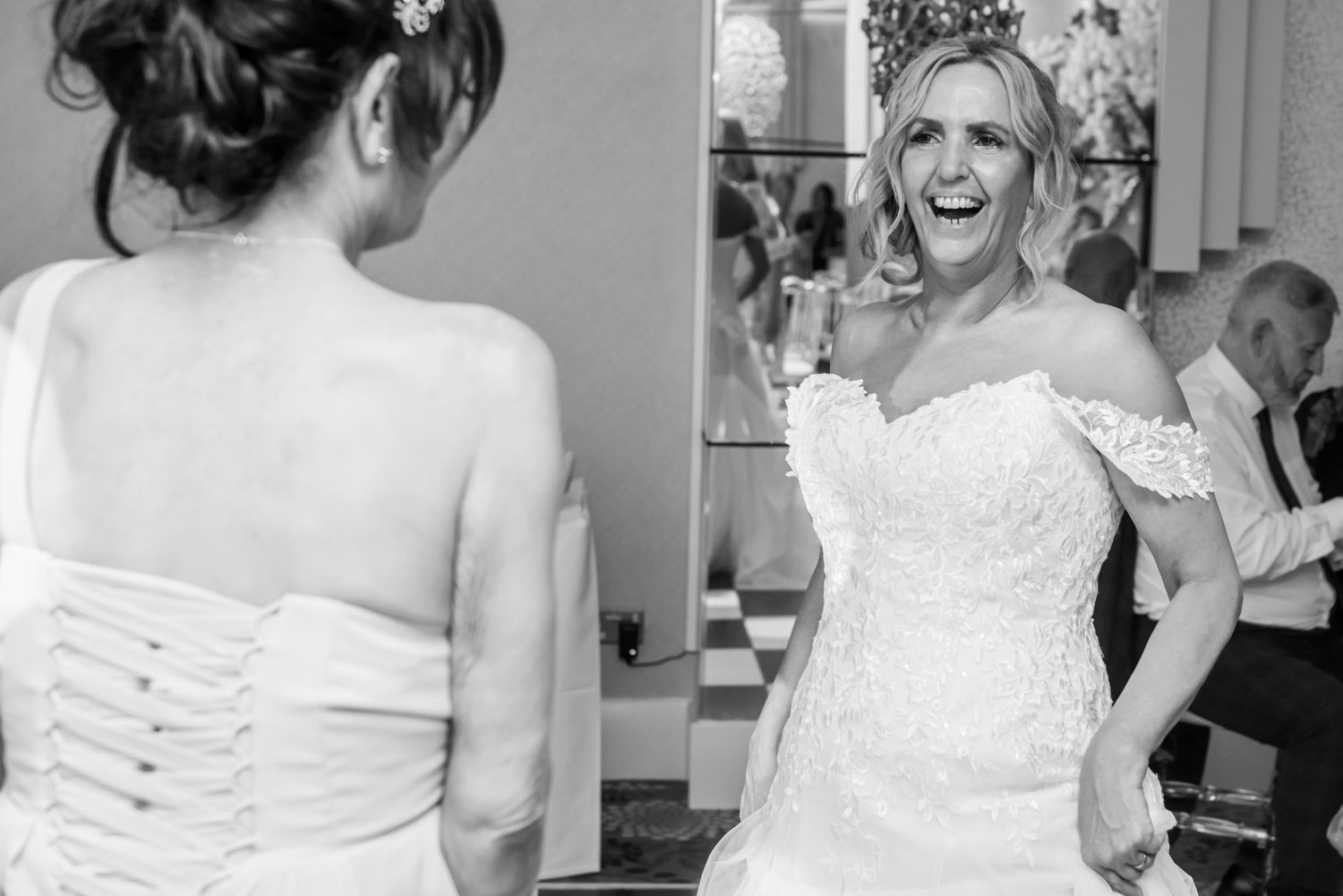 Black and white wedding photo showing bride in lace off-shoulder dress sharing joyful moment during reception.