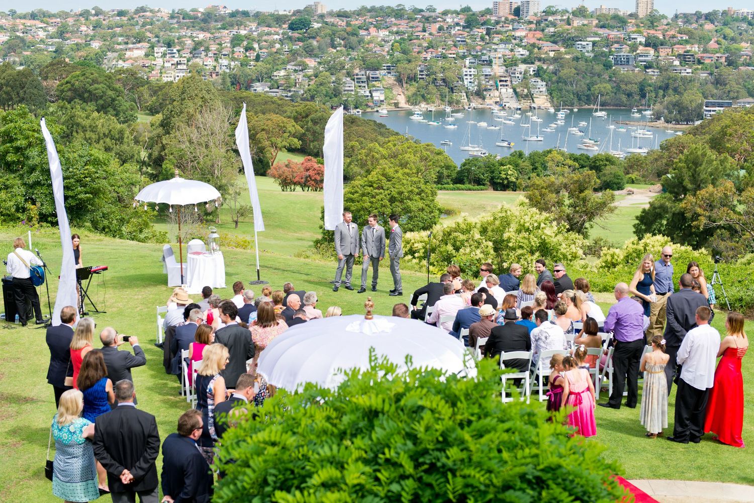 Scenic outdoor wedding ceremony overlooking a harbor with guests gathered on a grassy hillside in Sydney.