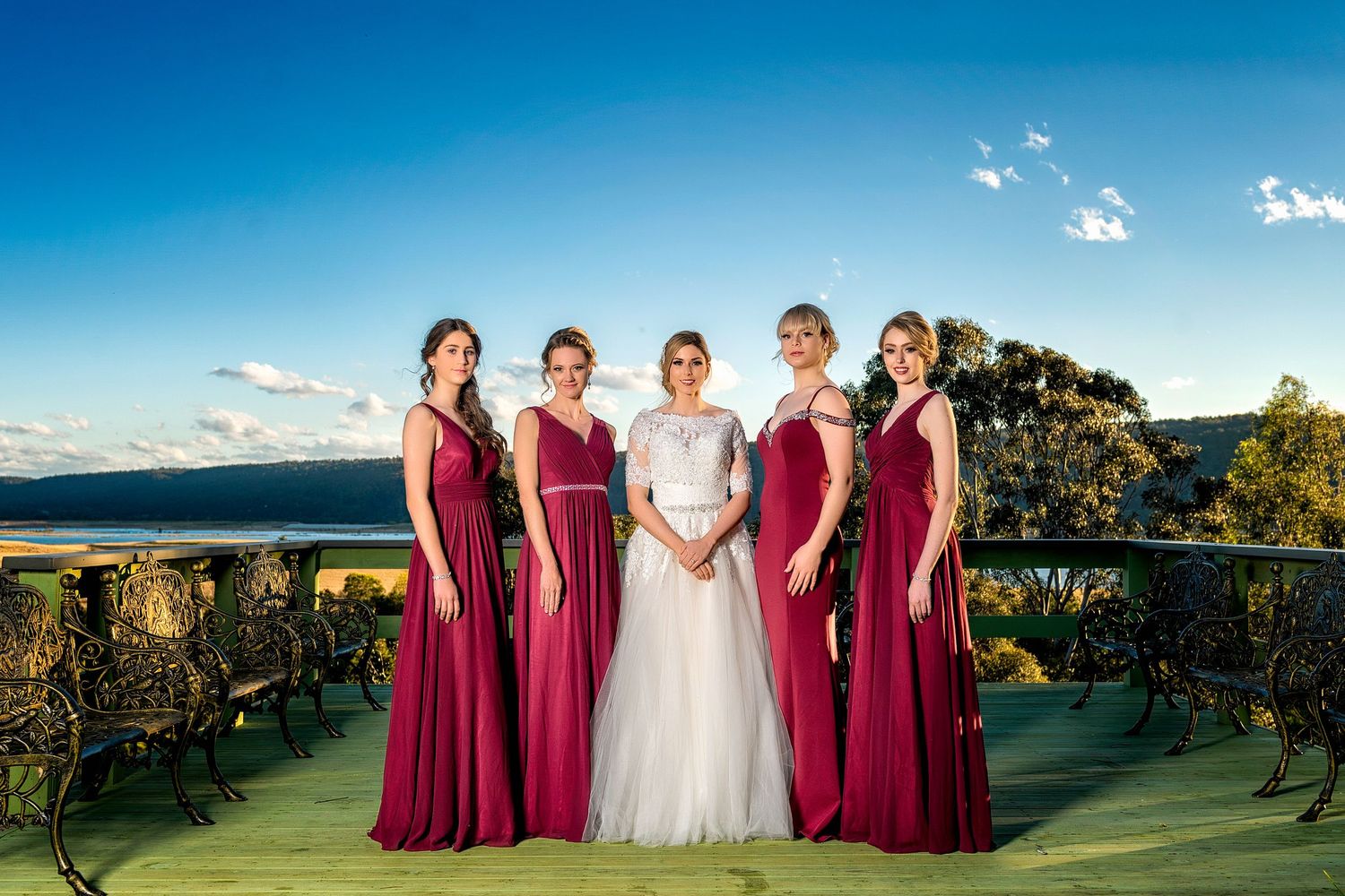 Bridesmaids in burgundy dresses and bride in white gown pose together on a scenic hilltop overlooking vineyards at sunset.