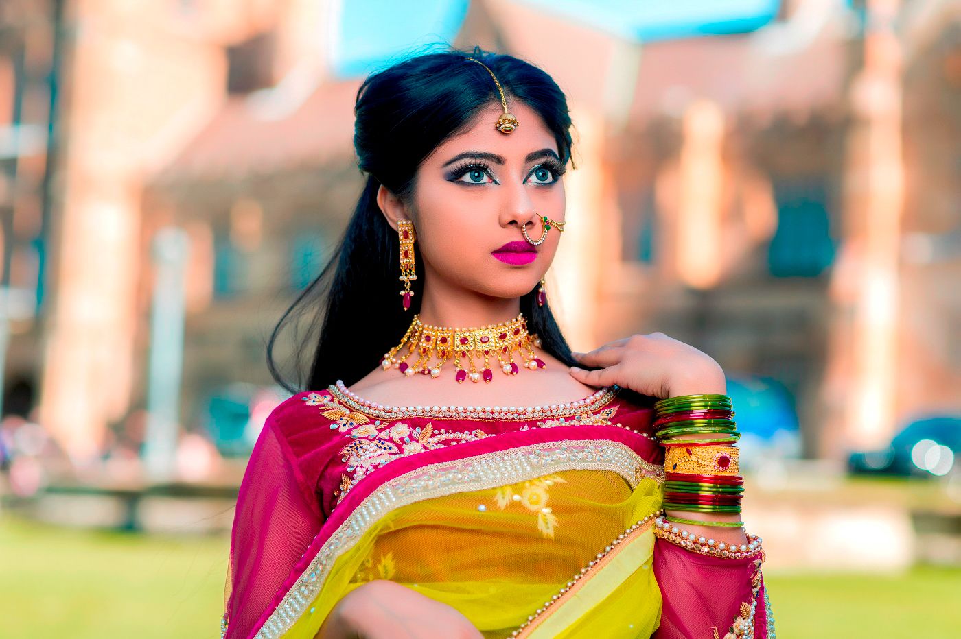 Woman in traditional Indian attire with yellow saree and pink blouse poses elegantly with jewelry in outdoor setting.