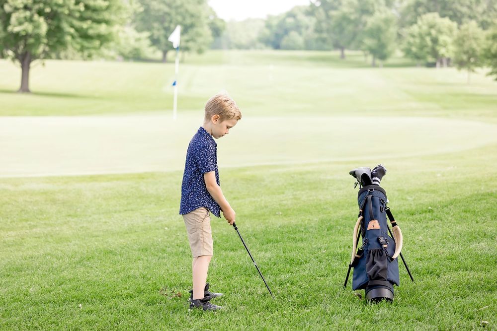 Young boy practicing golf on a lush green course, standing beside a junior golf bag with clubs.