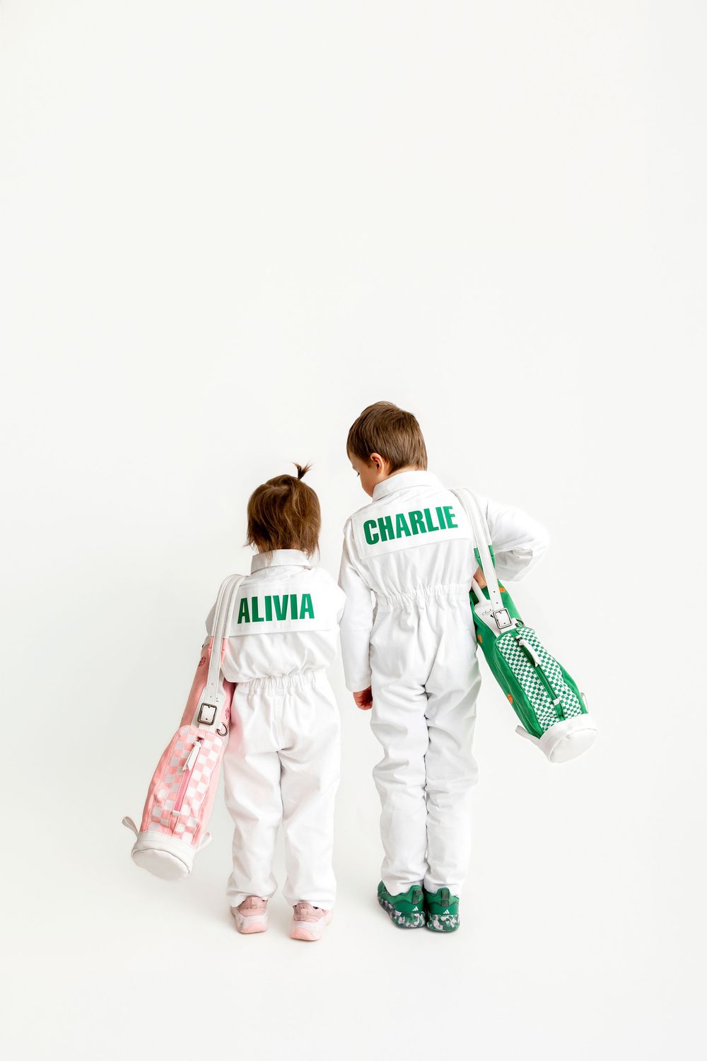 Two children in white jumpsuits named Alivia and Charlie hold golf bags, viewed from behind on white background.