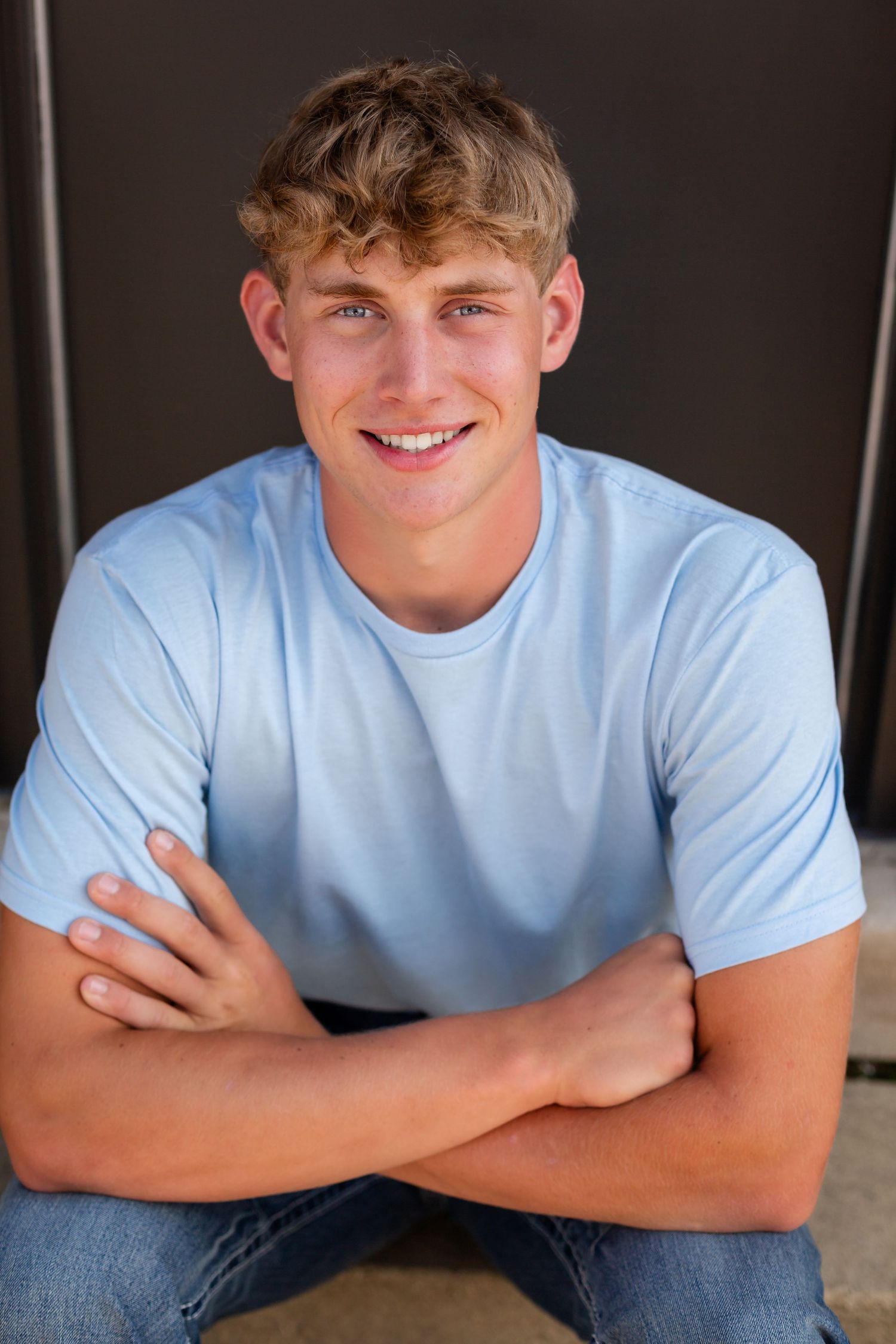 A young person with wavy blonde hair wearing a light blue t-shirt sits casually with crossed arms and a friendly expression.