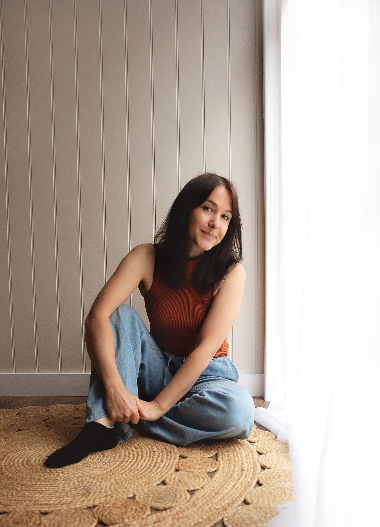 Jo Cove photography in rust-colored sleeveless top and jeans sits on round jute rug by bright window in newborn photography studio