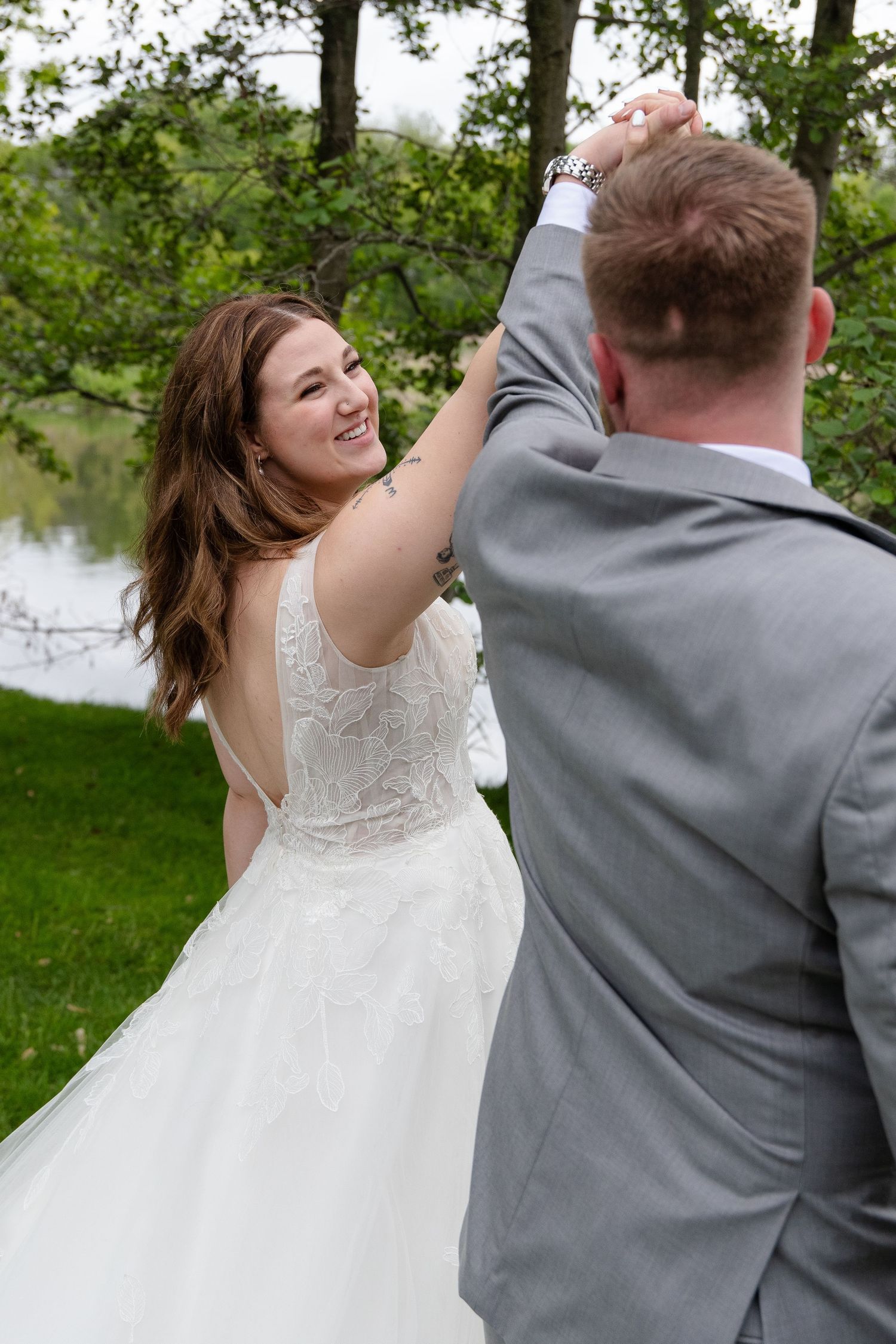 A couple in wedding attire dancing together outdoors near a lake with trees in the background.