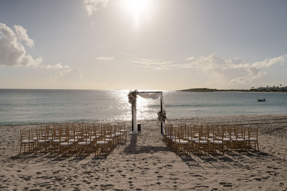 Beach wedding ceremony setup with wooden chairs and floral arch on sandy shore at sunset overlooking ocean.