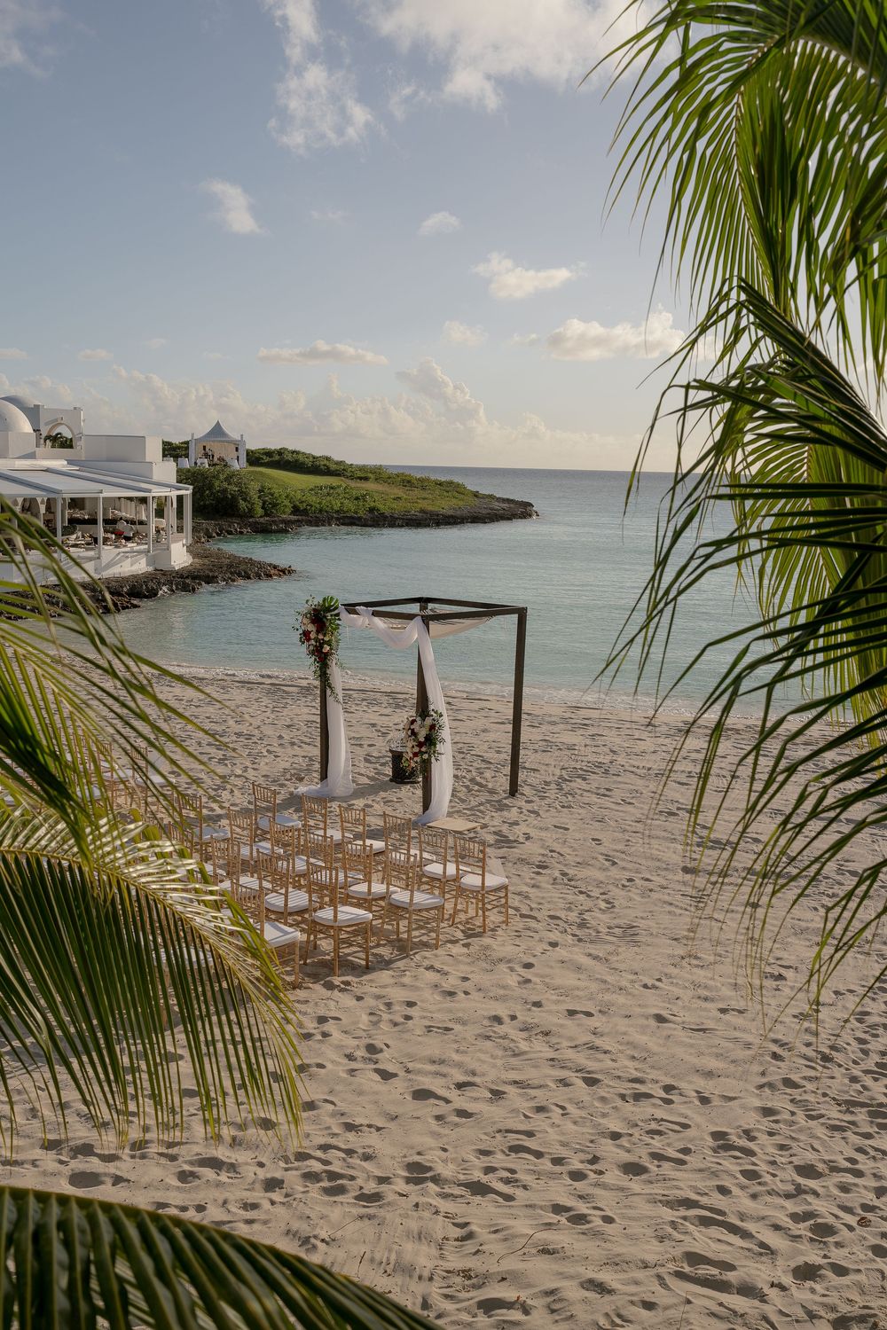 Rows of white chairs arranged on a tropical beach with palm trees and an arch setup awaiting a wedding ceremony.