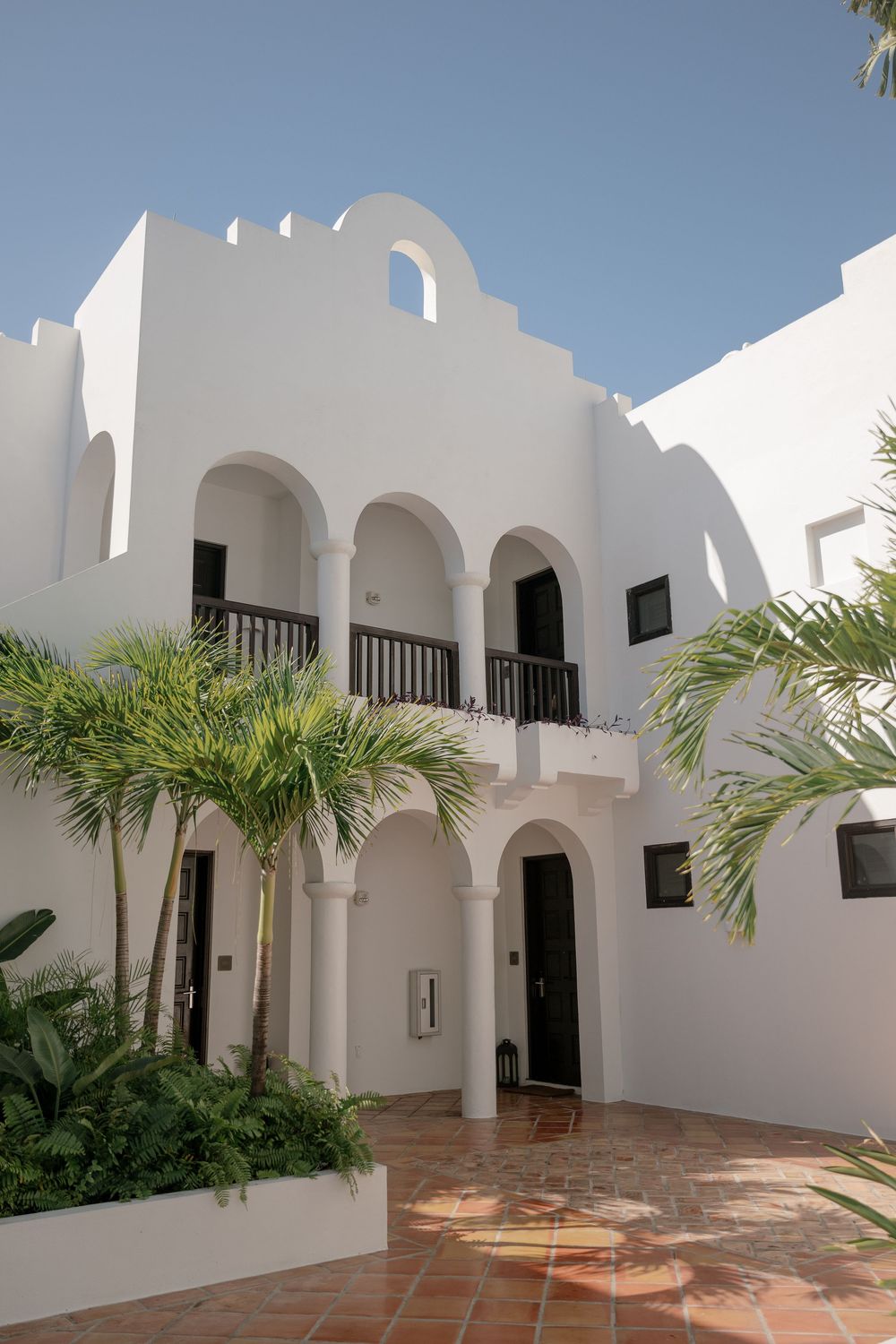 Mediterranean-style white villa with arched doorways, balconies and palm trees under blue sky.