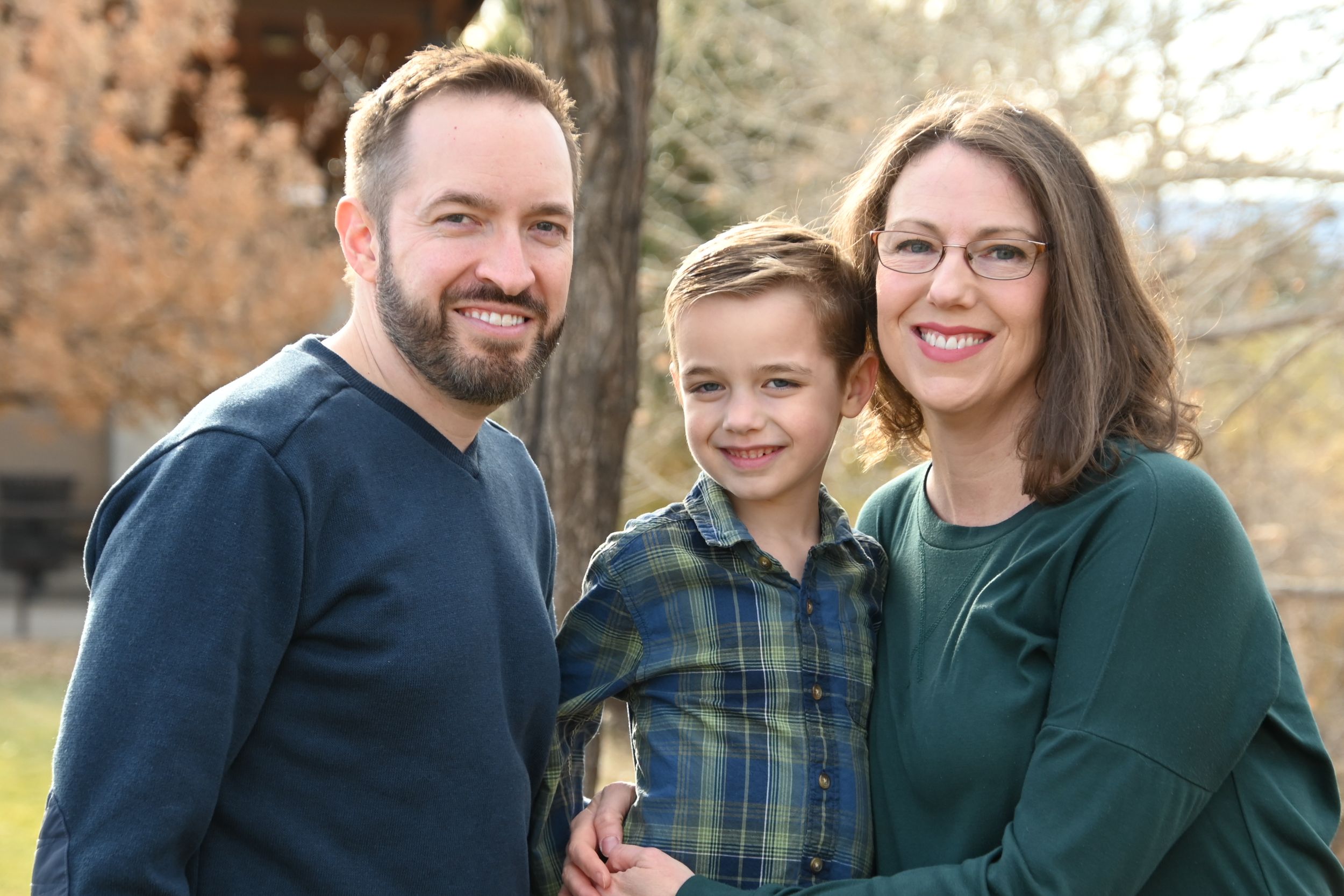 A family of three wearing casual clothing poses together outdoors on an autumn day.
