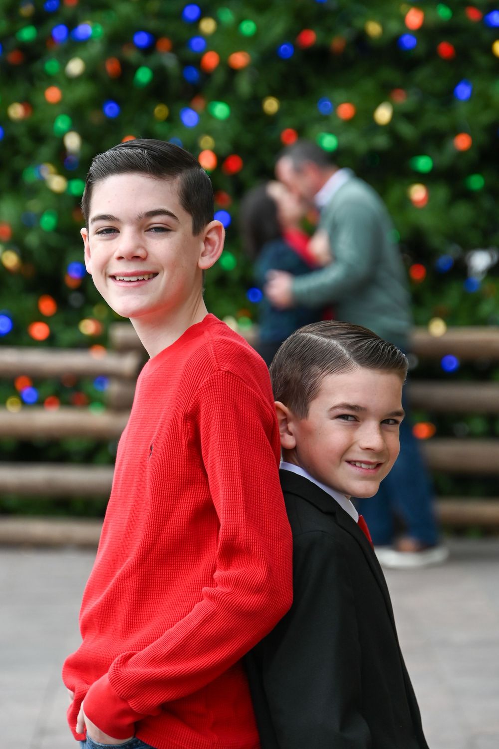 Two children wearing red and black sweaters pose together against a festive holiday backdrop with colorful lights.