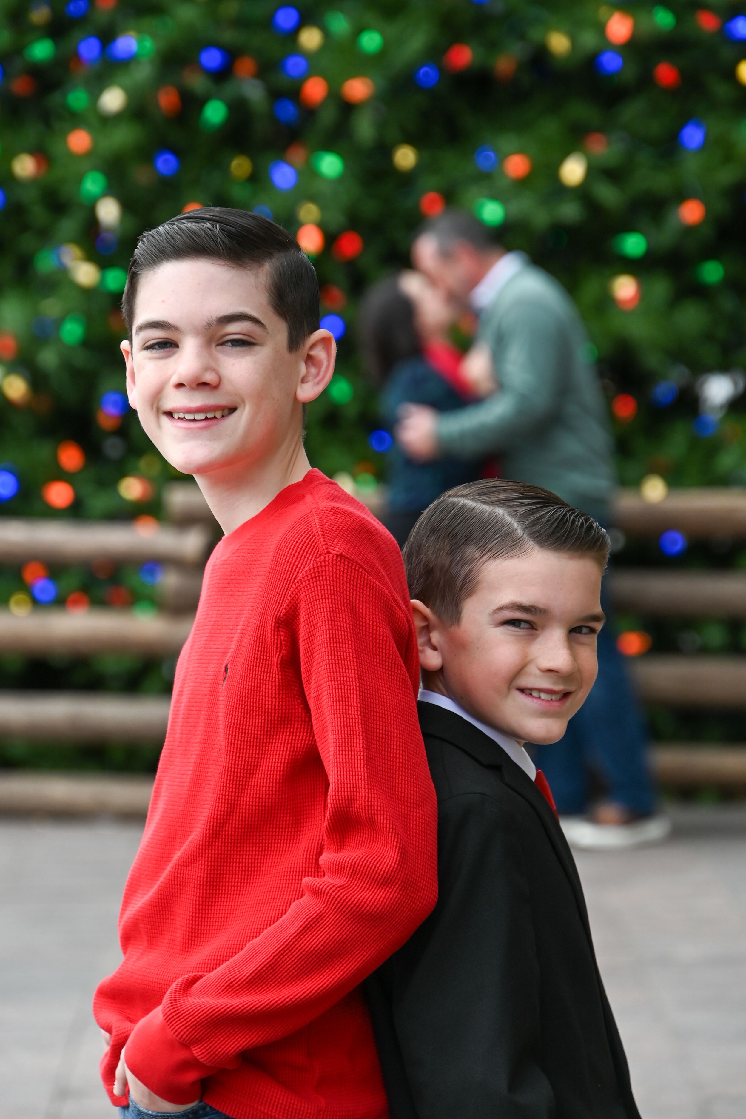 Two children wearing red and black sweaters pose together against a festive holiday backdrop with colorful lights.
