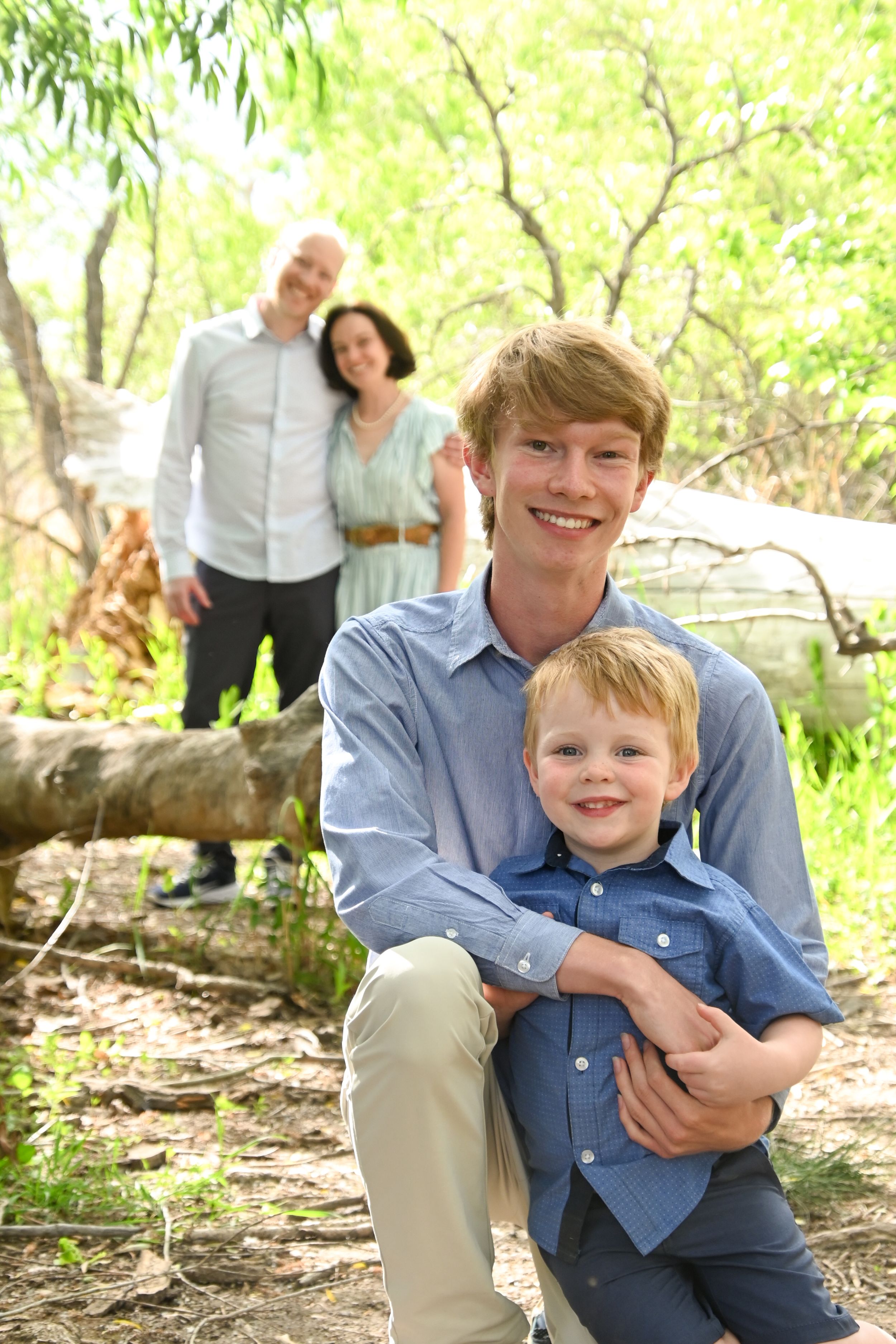 Family enjoying time together outdoors on a sunny day in a natural wooded setting with fallen logs and green foliage.