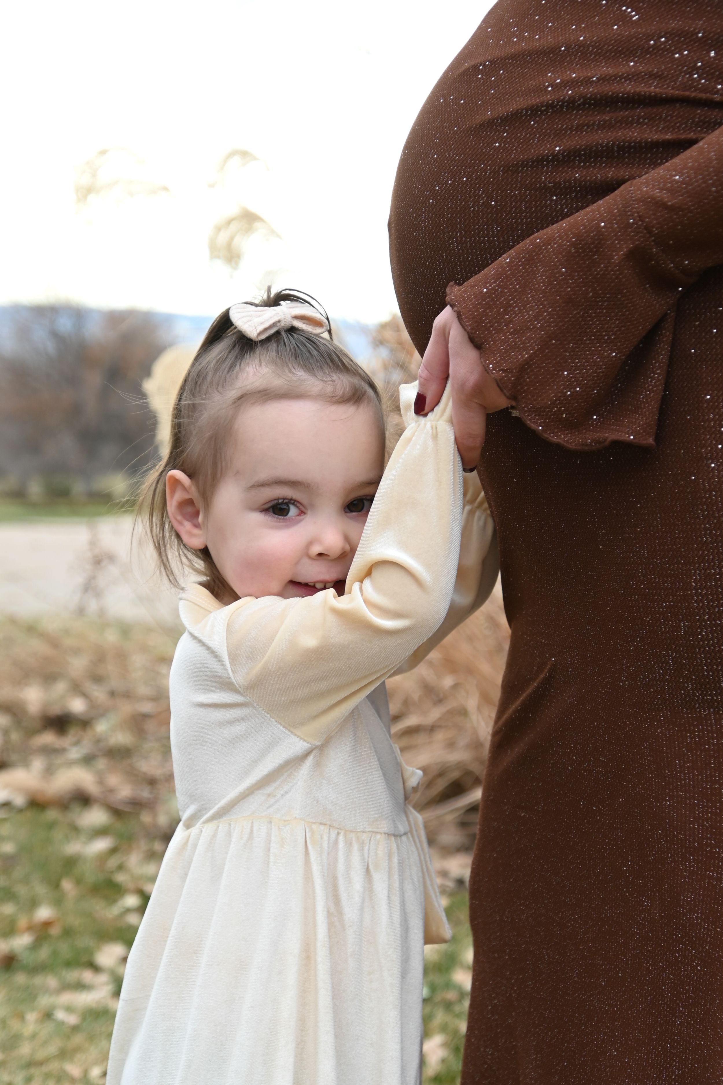 Small child in cream-colored dress gently holding onto a person wearing brown clothing outdoors.