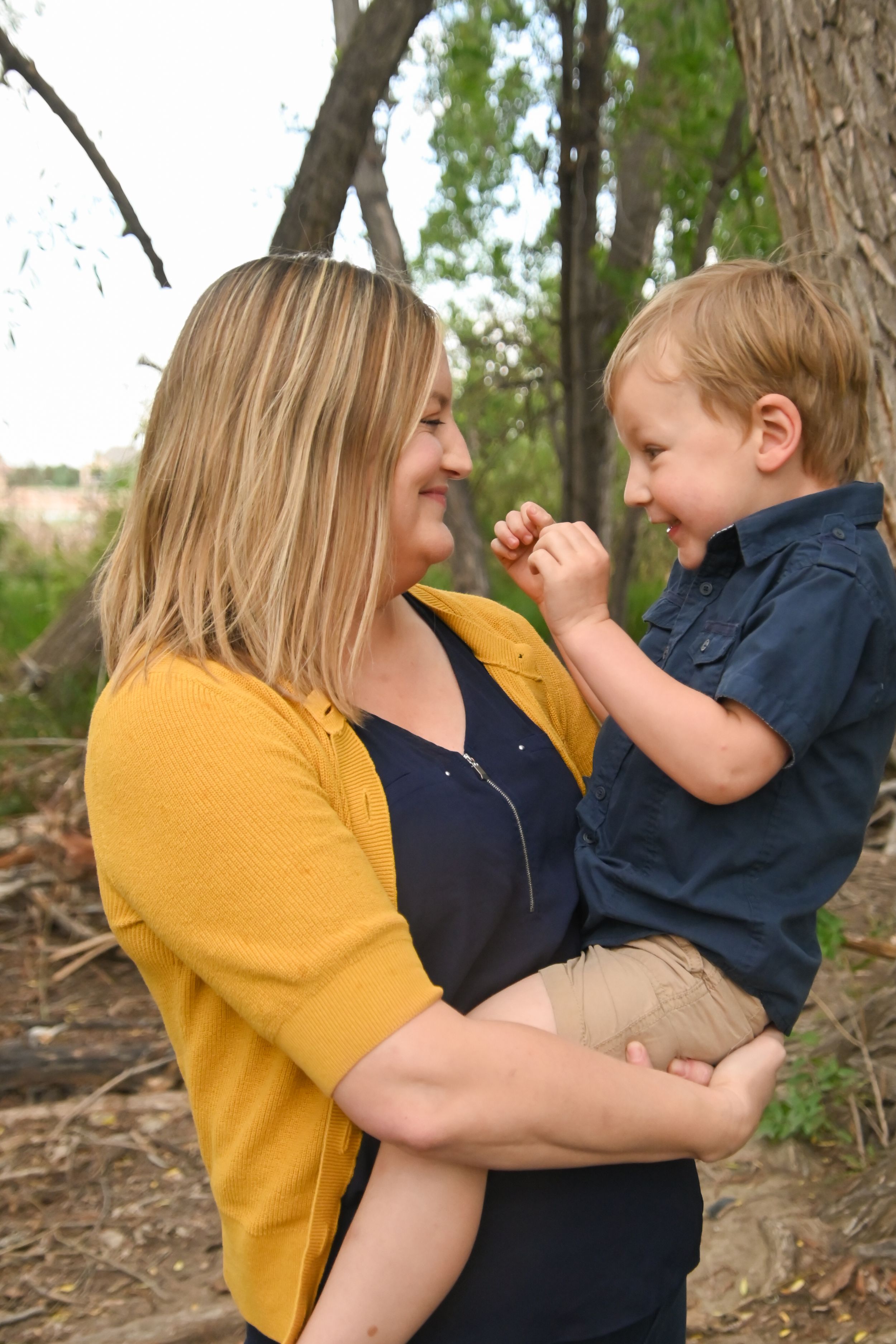 A parent in a yellow cardigan holds a laughing toddler in a navy shirt outdoors among trees.