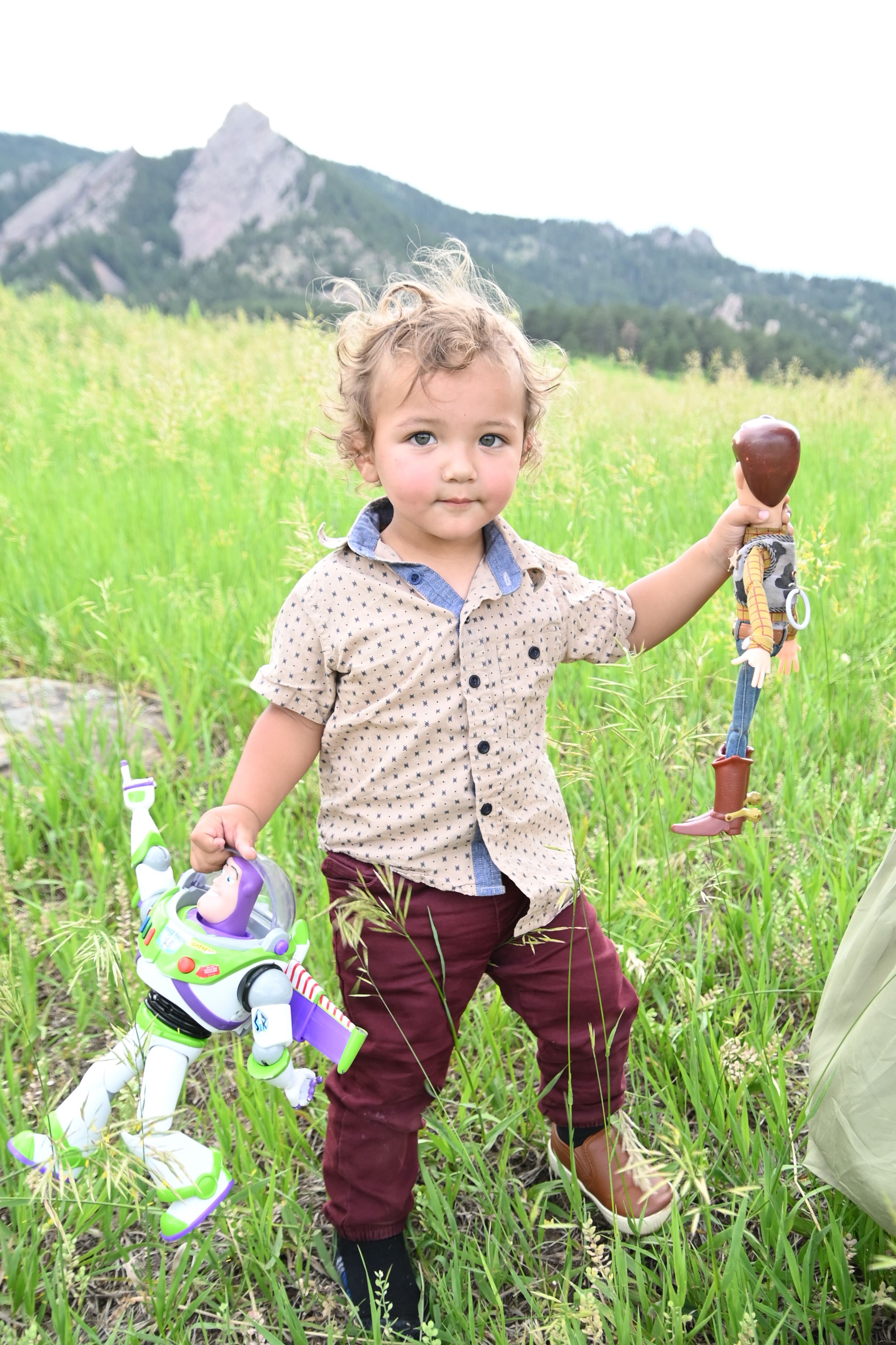 Child plays joyfully in a green meadow with mountain backdrop while wearing burgundy pants and floral shirt.