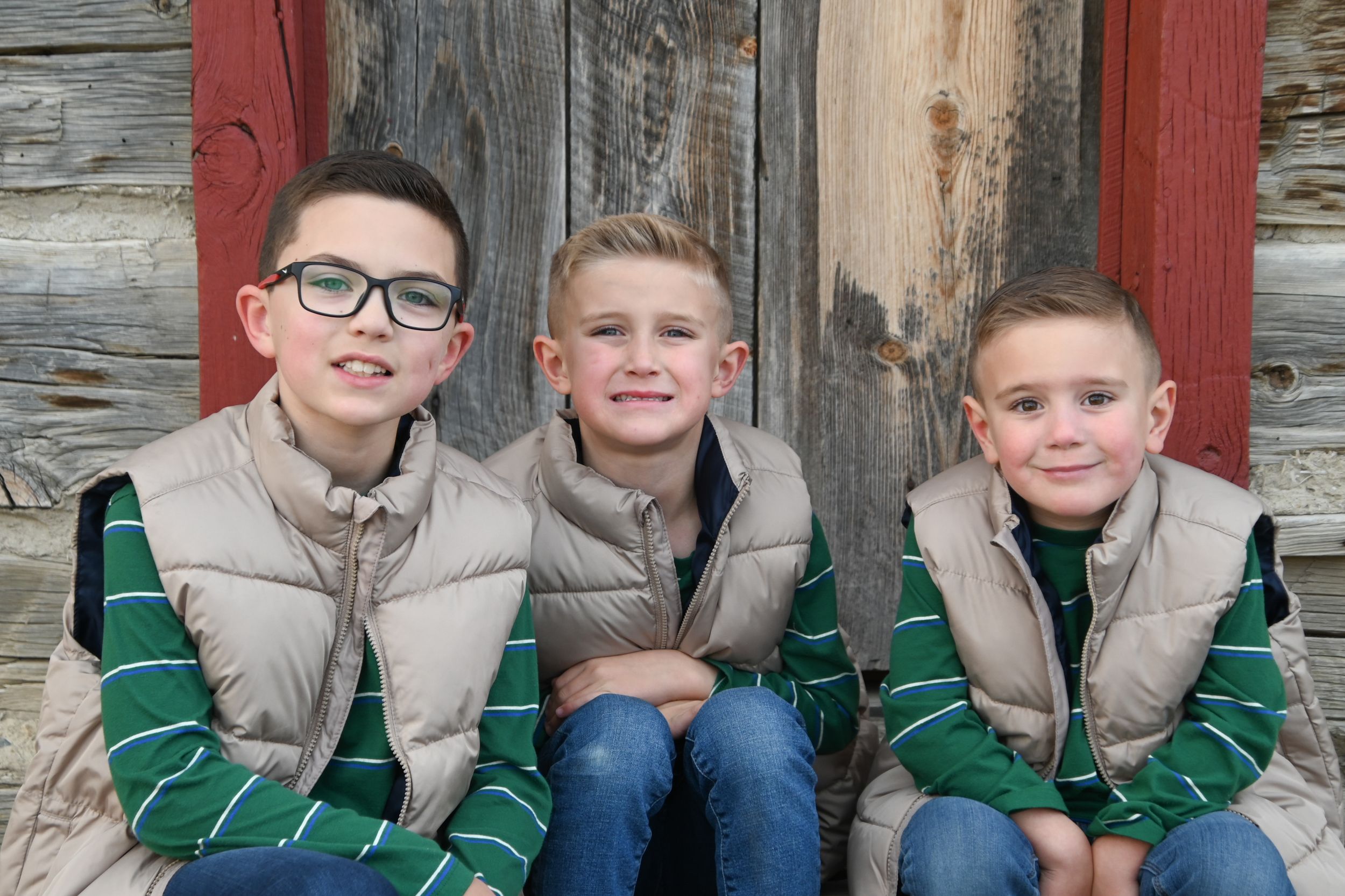 Three young boys in matching beige vests and green striped shirts sit together on rustic wooden steps.