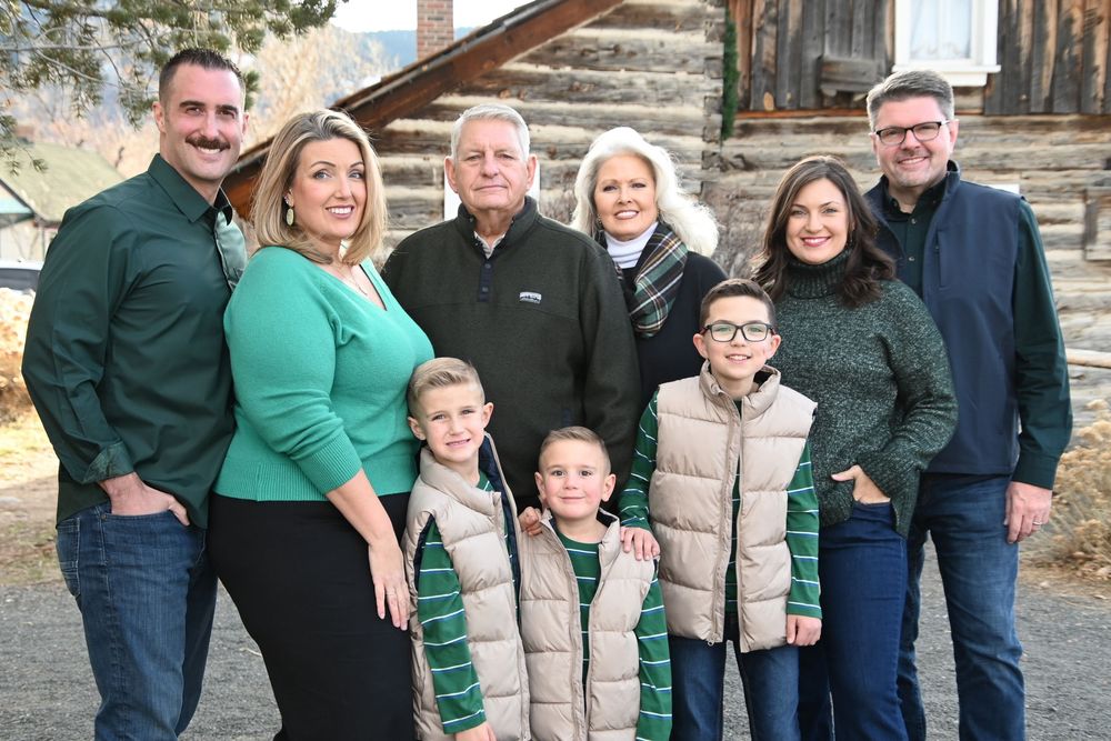 A family group wearing coordinated green and beige outfits poses together outdoors in front of a rustic building.