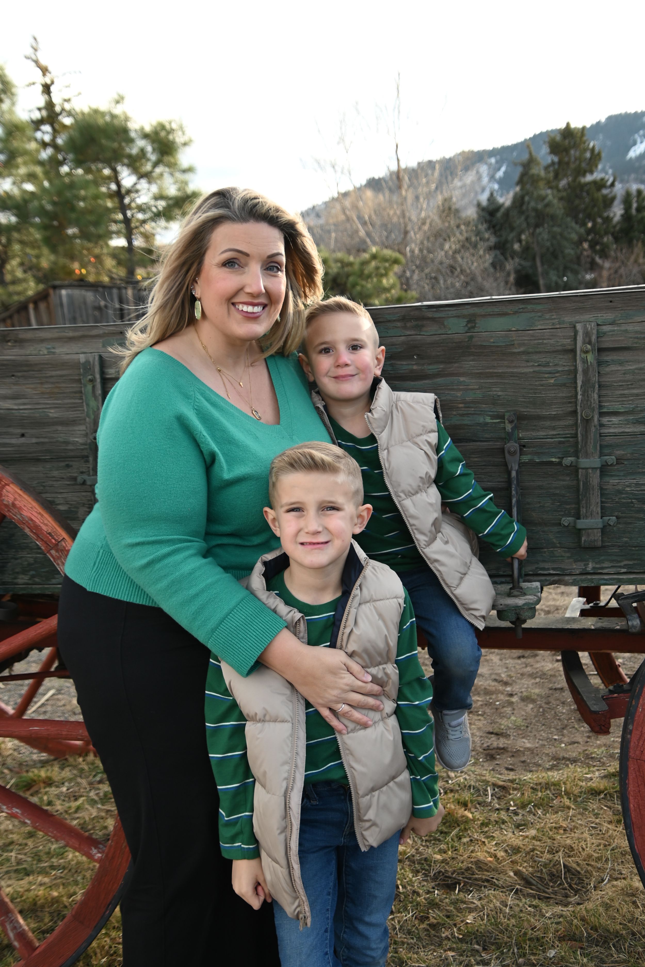A small family poses together in front of an old wooden wagon while wearing coordinated green and beige winter outfits.