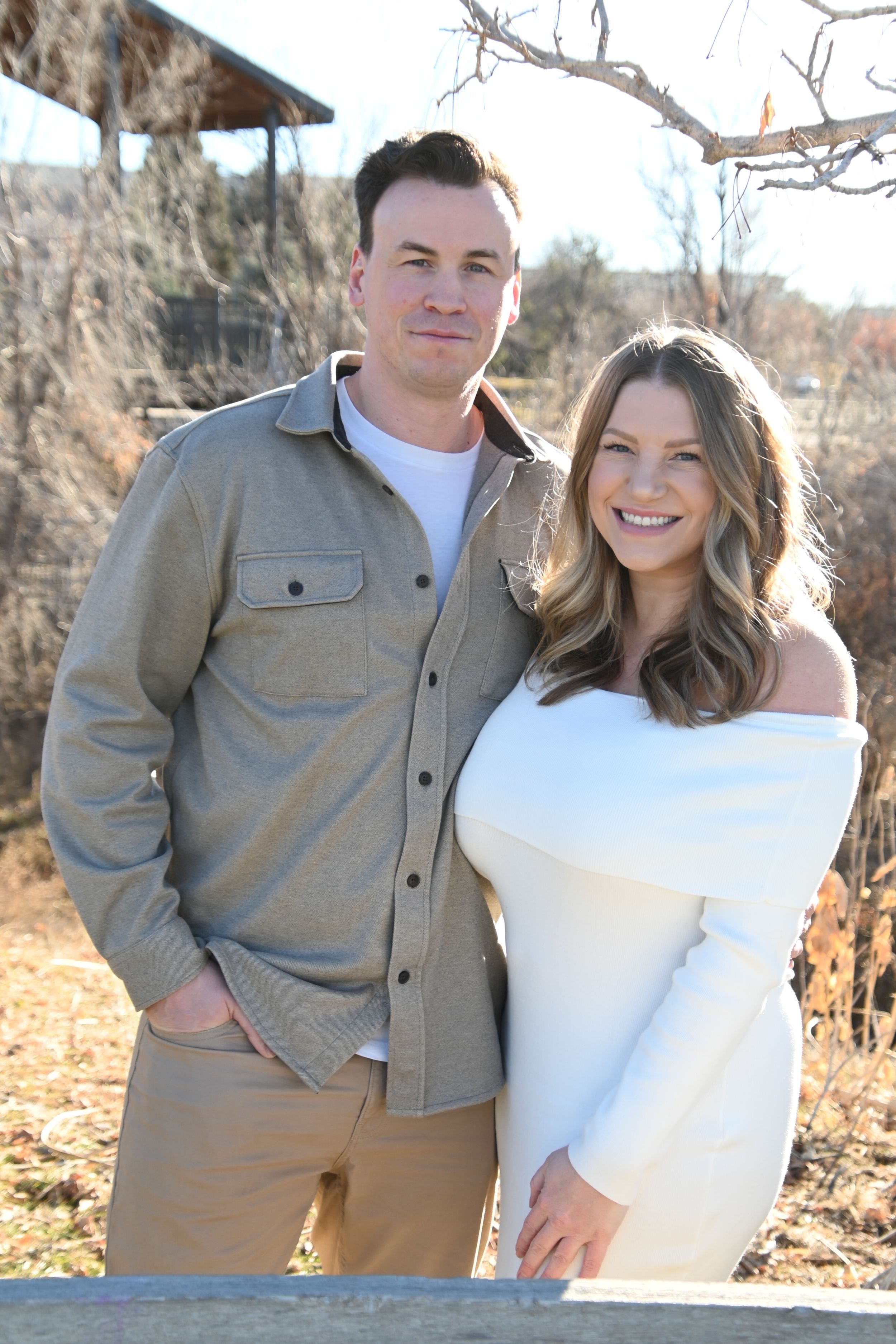 A couple poses together outdoors in neutral clothing during an autumn photoshoot with bare branches in the background.